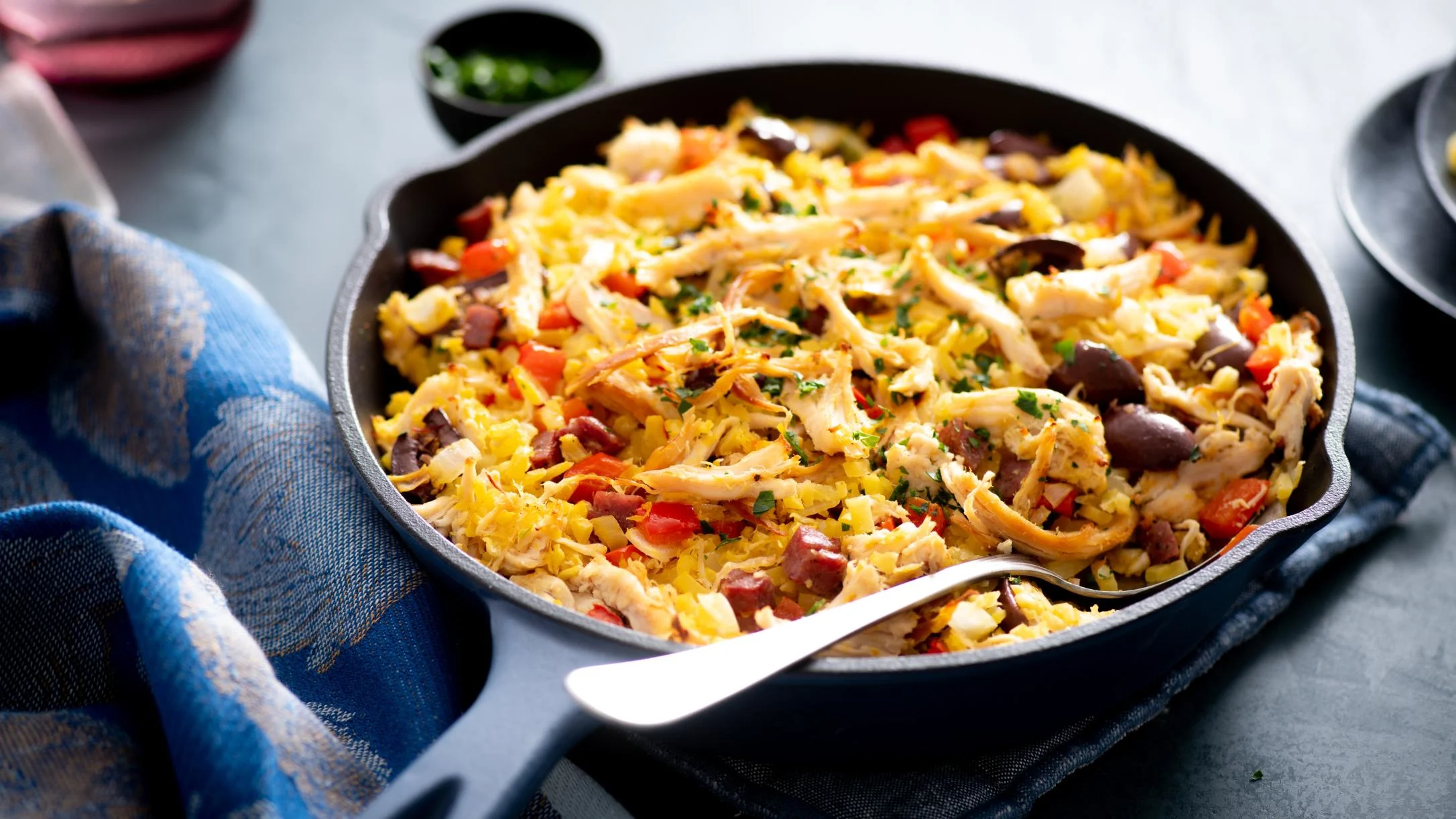 A cast iron skillet filled with shredded chicken, yellow cauliflower rice, red peppers, olives, and herbs, with a spoon resting inside.