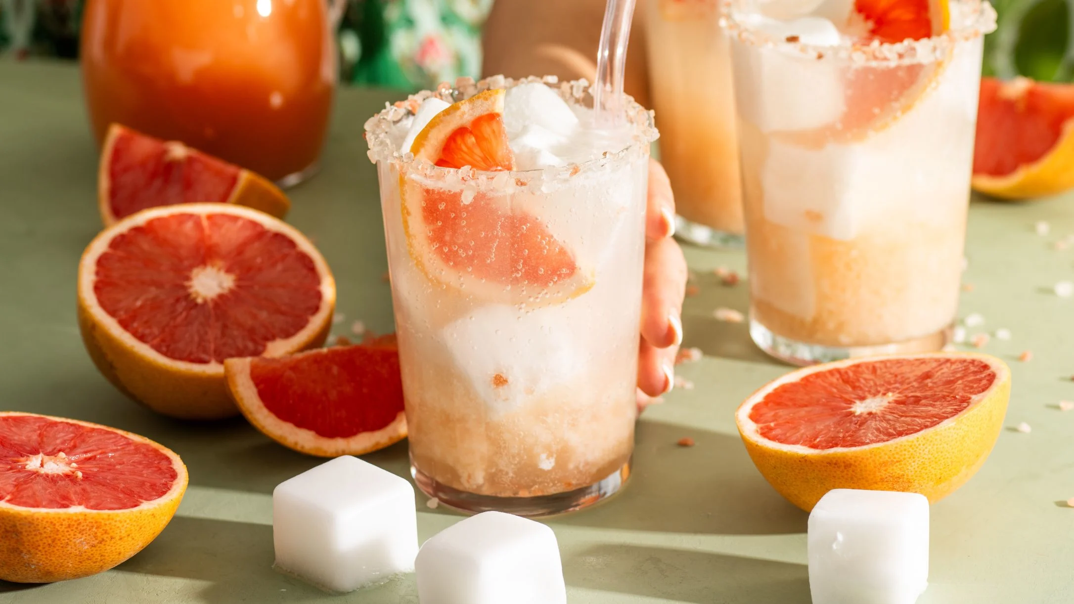 Grapefruit cocktails with salt rims and fresh fruit slices, surrounded by halved grapefruits and sugar cubes on a green surface.