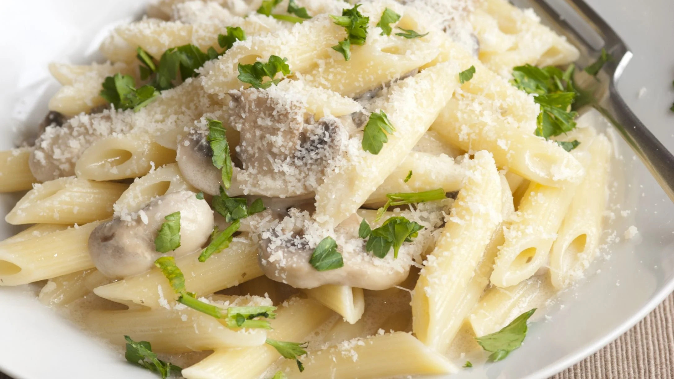 A white plate of penne pasta with mushrooms, topped with grated parmesan and fresh chopped parsley, with a fork nearby.