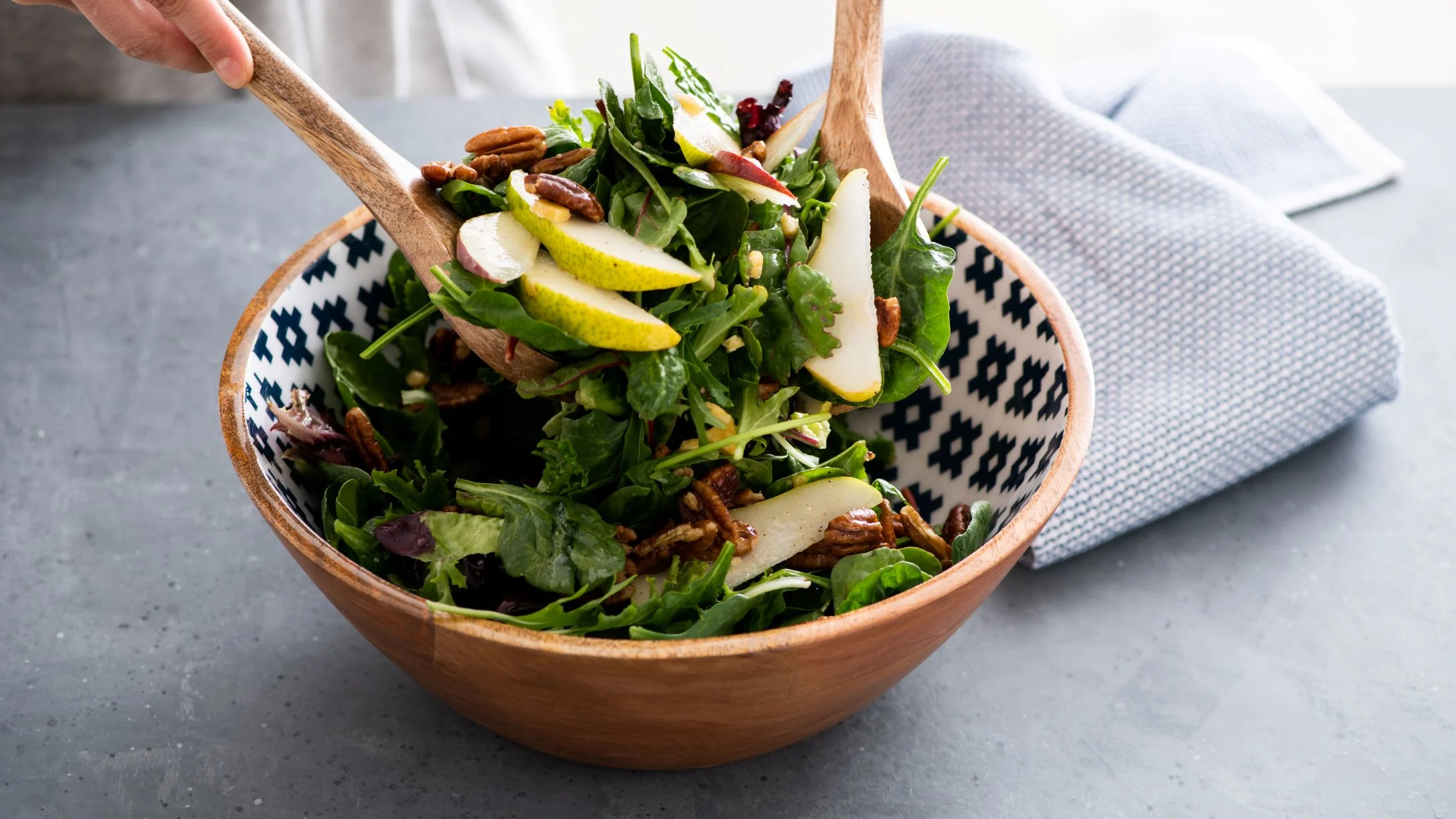 Fresh green salad with pear slices and pecans being tossed in a decorative wooden bowl with serving utensils.