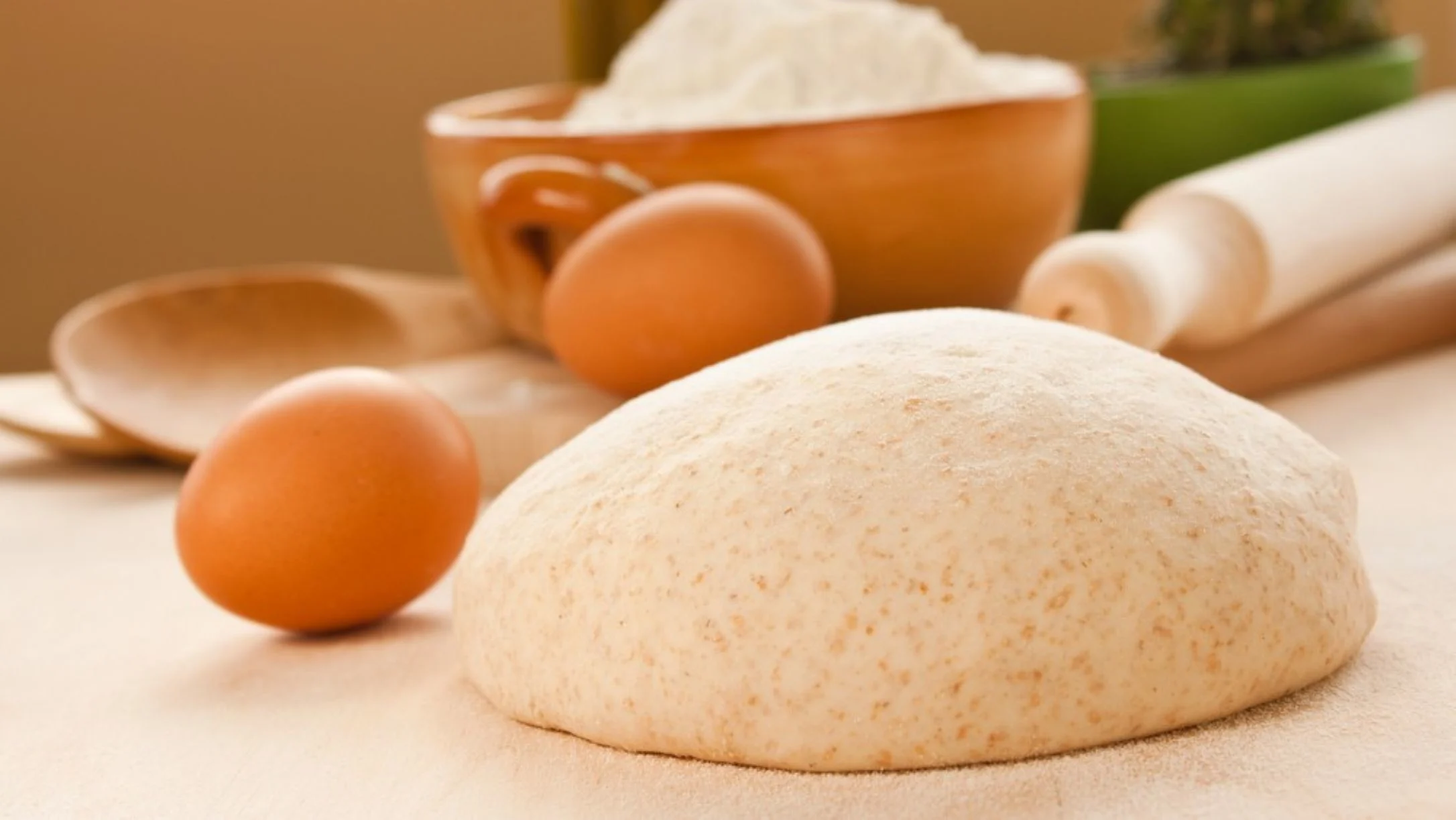 A round ball of pizza crust dough on a floured surface with eggs, a bowl of flour, wooden spoon, and rolling pin nearby.