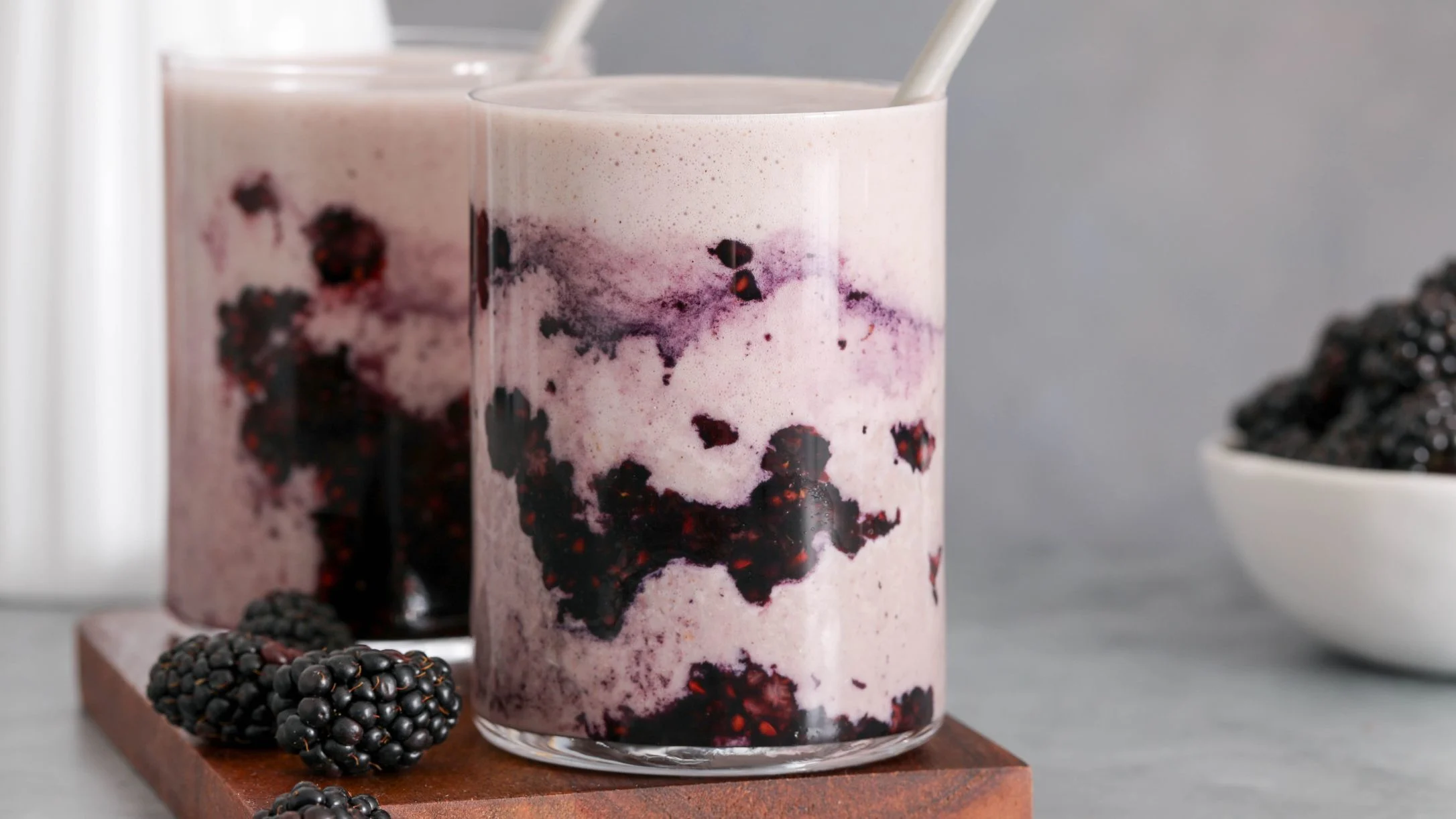 Two glasses of creamy blackberry smoothie with fresh blackberries on a wooden board, and a bowl of blackberries nearby.