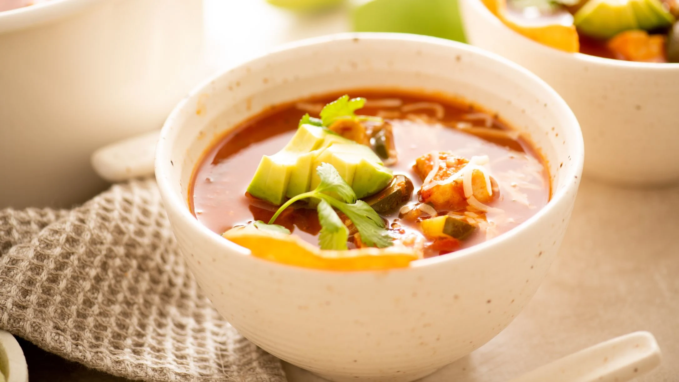 White bowl of hearty vegetable soup with avocado and herbs garnish, served on textured cloth.