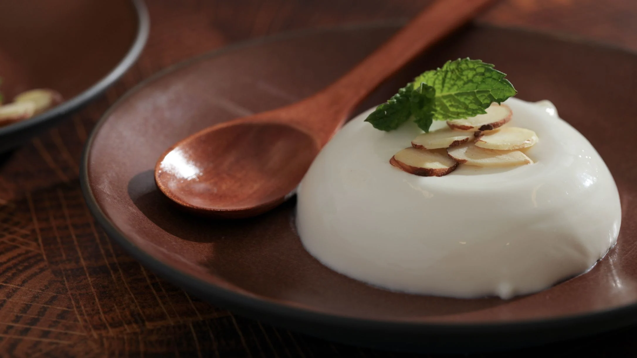 White panna cotta topped with sliced almonds and fresh mint on a dark plate with a wooden spoon.