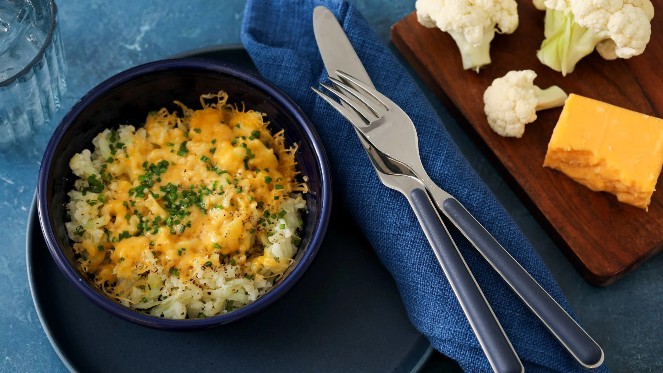 Cauliflower mash bowl topped with melted cheese and chives, served with utensils and raw cauliflower on wooden board.