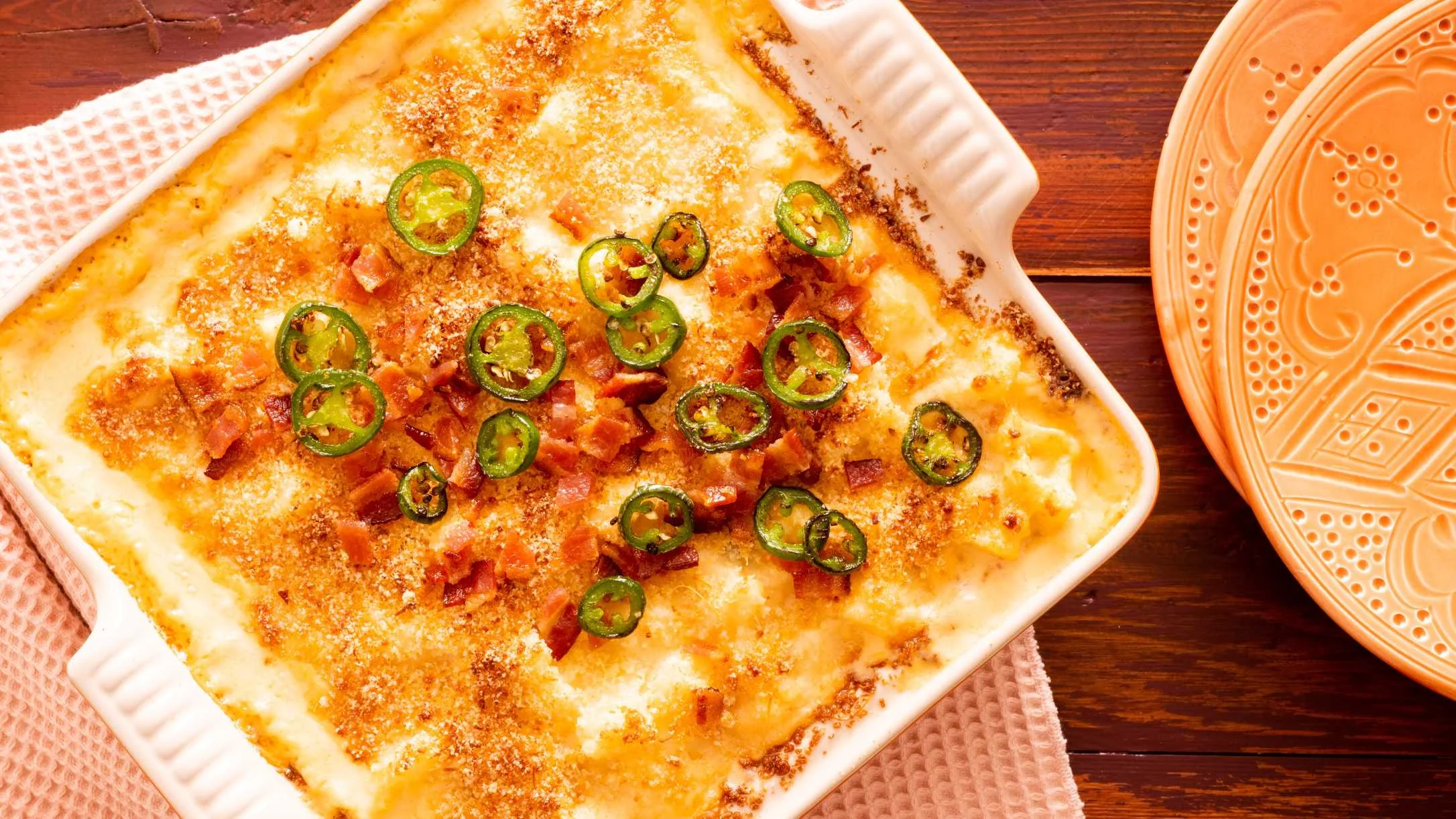 Cheesy baked casserole topped with sliced jalapeños and bacon bits in a white ceramic dish beside an orange plate.