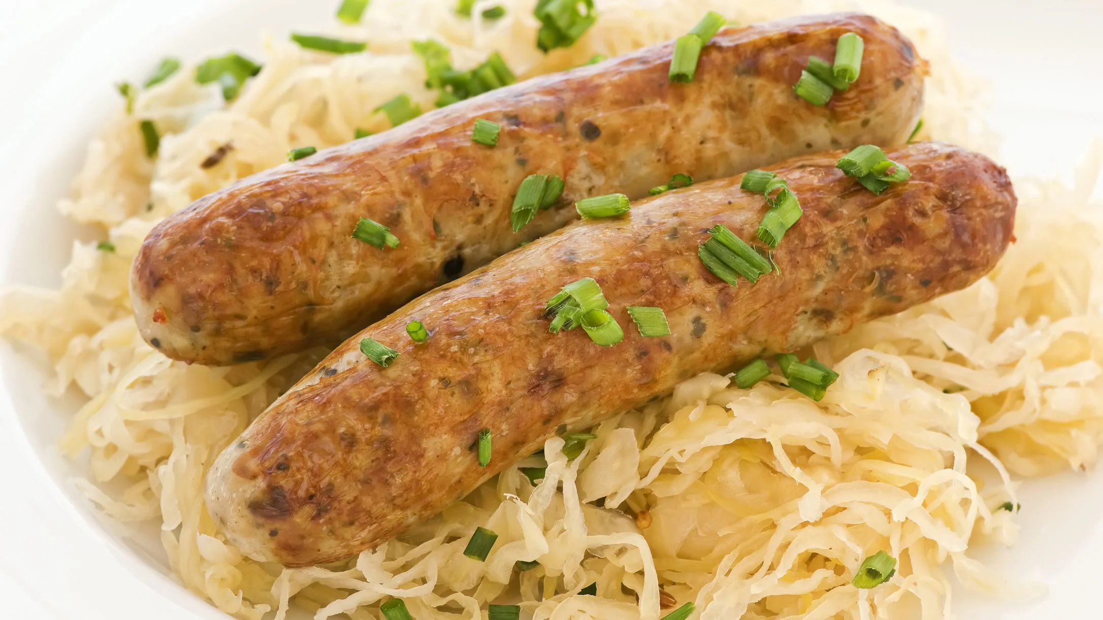 Two golden-brown grilled bratwurst topped with chopped chives, served on a bed of sauerkraut on a white plate.