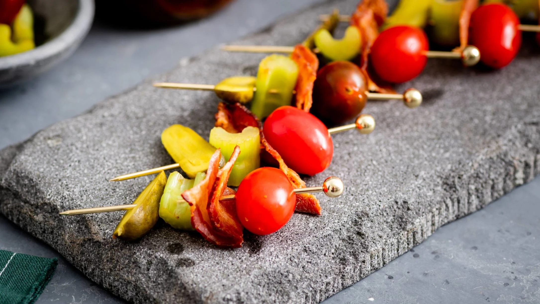 Appetizer skewers with cherry tomatoes, pickles, and bacon pieces arranged on a gray stone serving board.