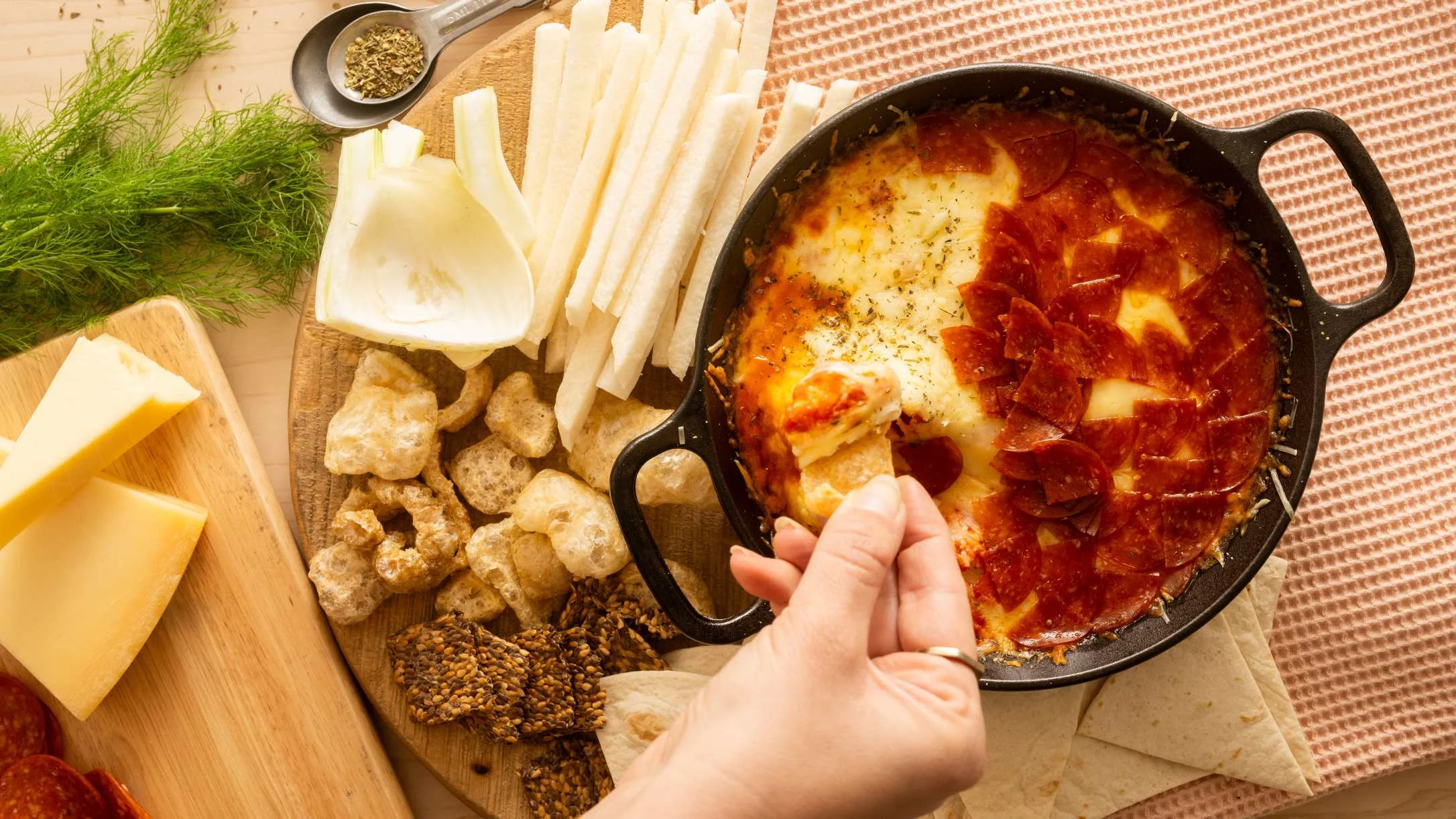 Hand dipping tortilla into pepperoni cheese dip in cast iron skillet, surrounded by jicama sticks, crackers, and fresh herbs.