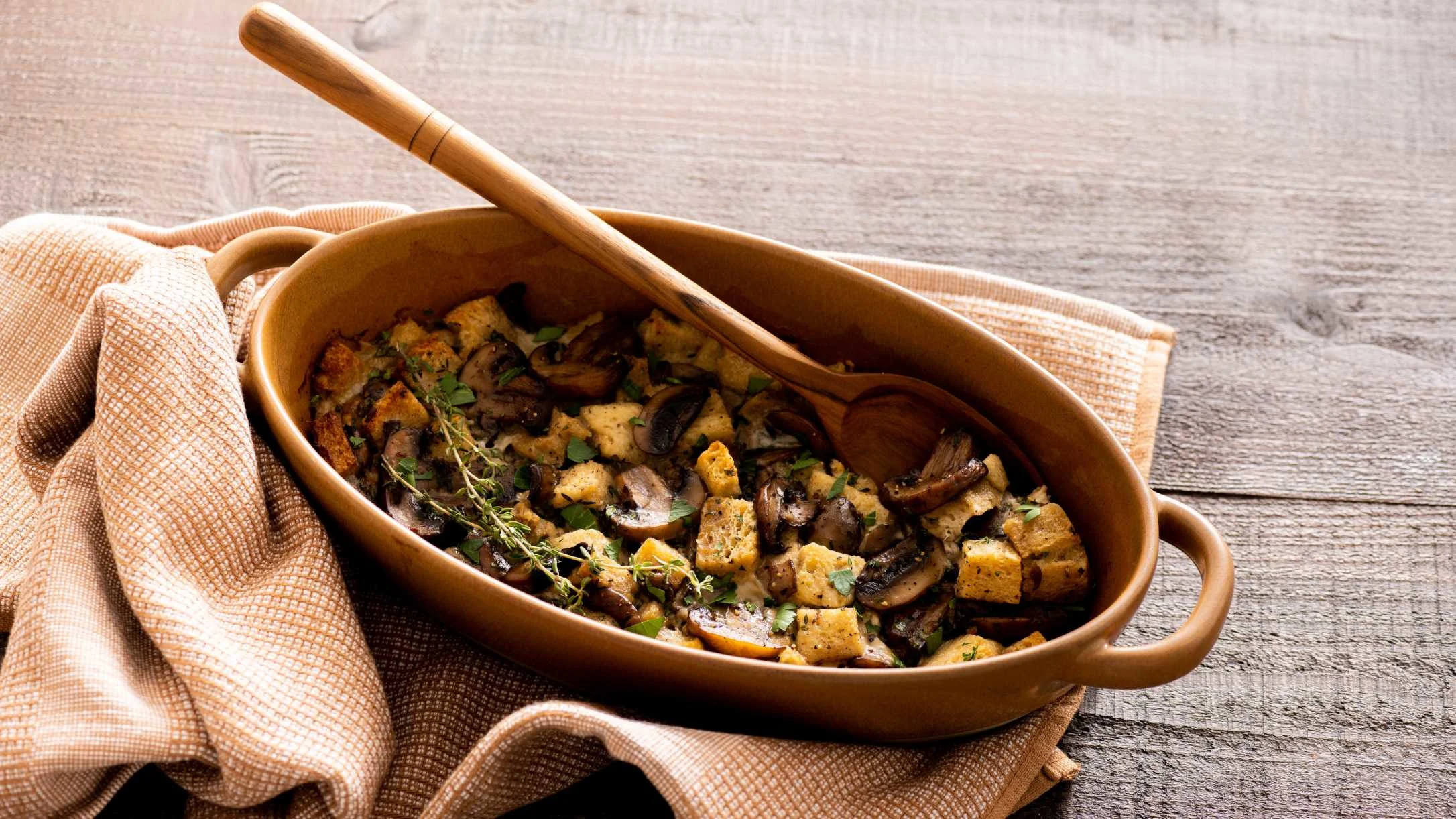 Rustic ceramic dish of mushroom and bread stuffing with wooden spoon, served on beige napkin against wooden surface.