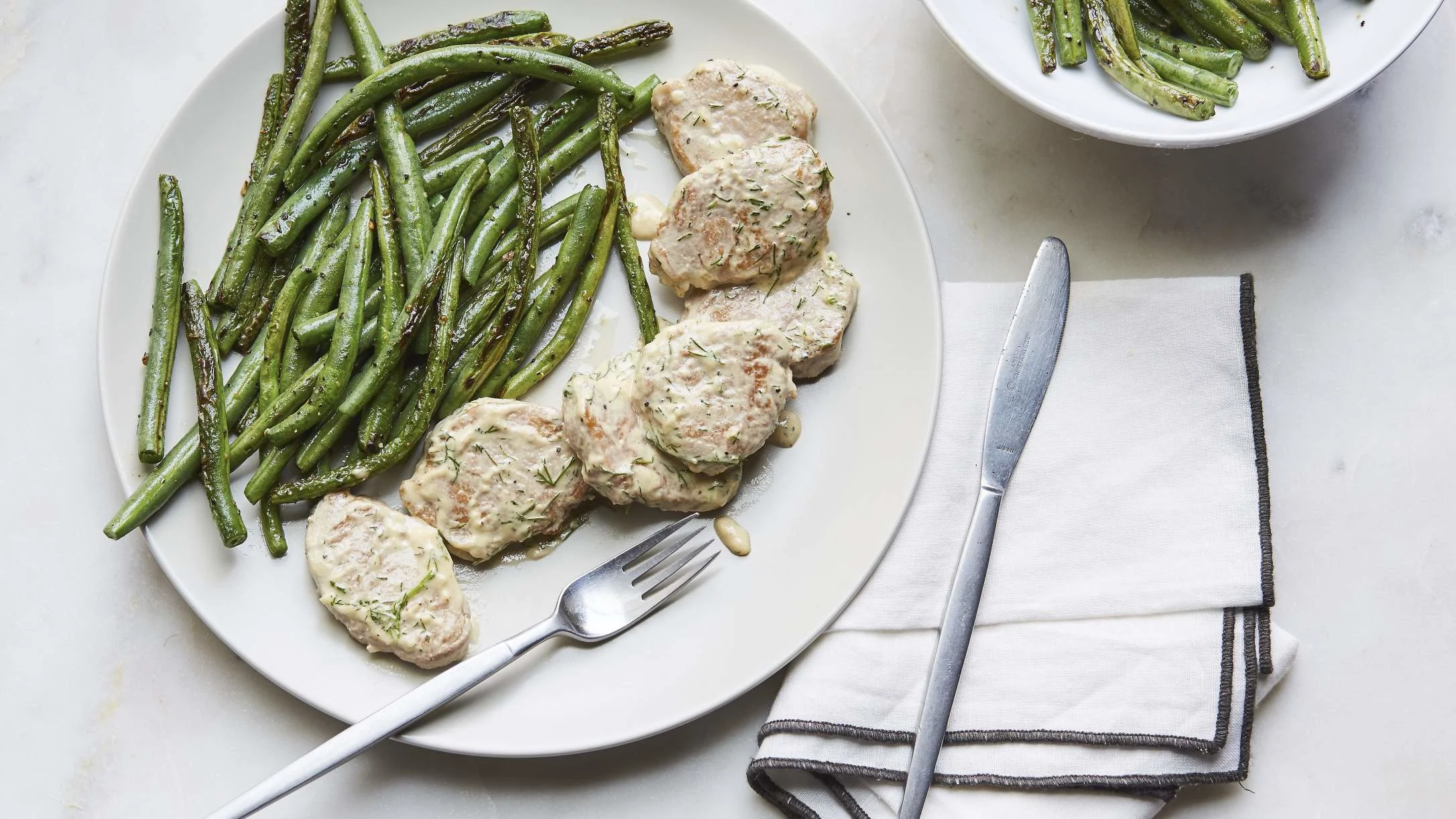 White plate with creamy pork medallions and roasted green beans, fork on plate, knife and napkin beside it.