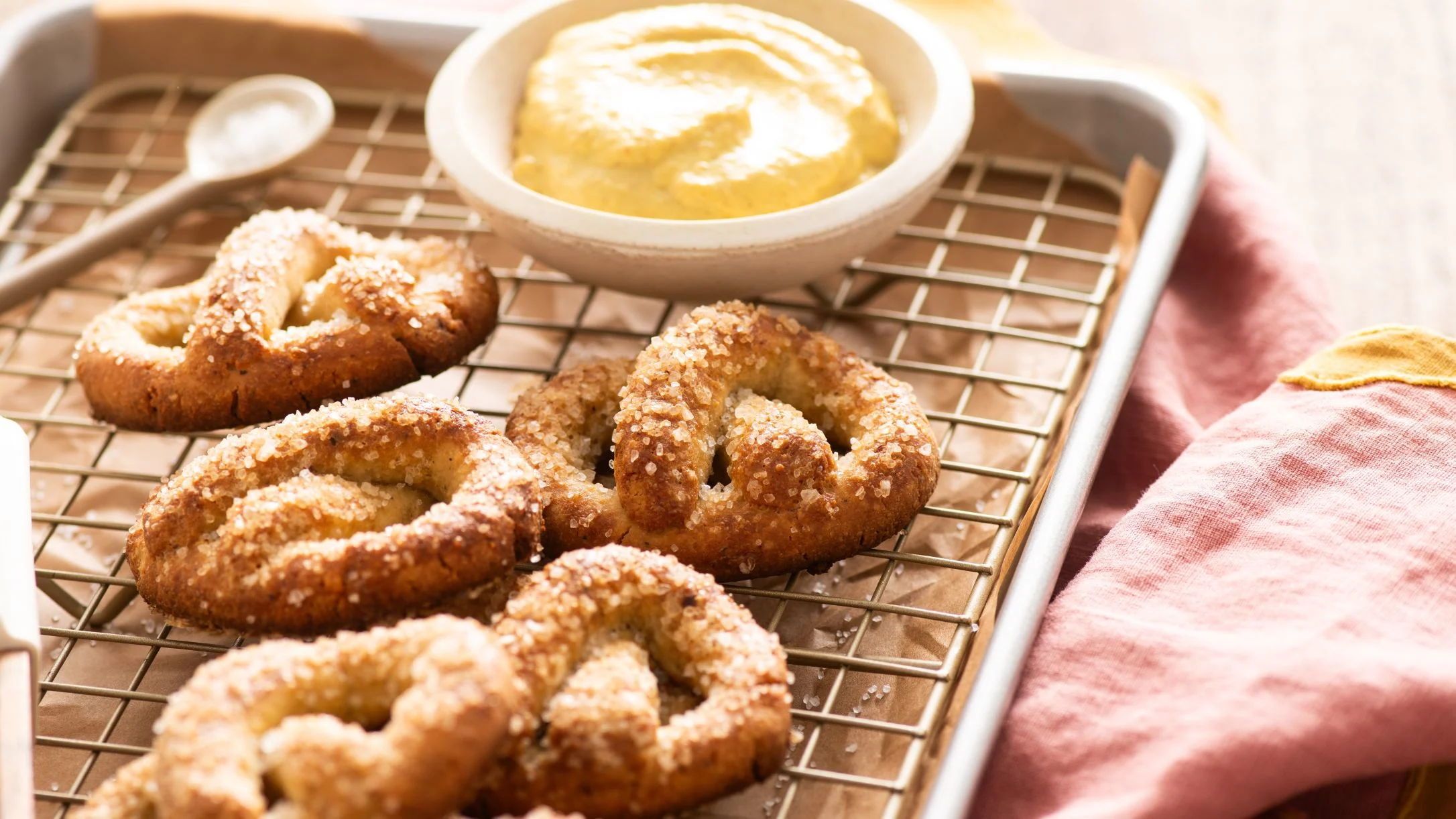 Golden-brown salted soft pretzels on a wire cooling rack with a bowl of yellow mustard dipping sauce nearby.