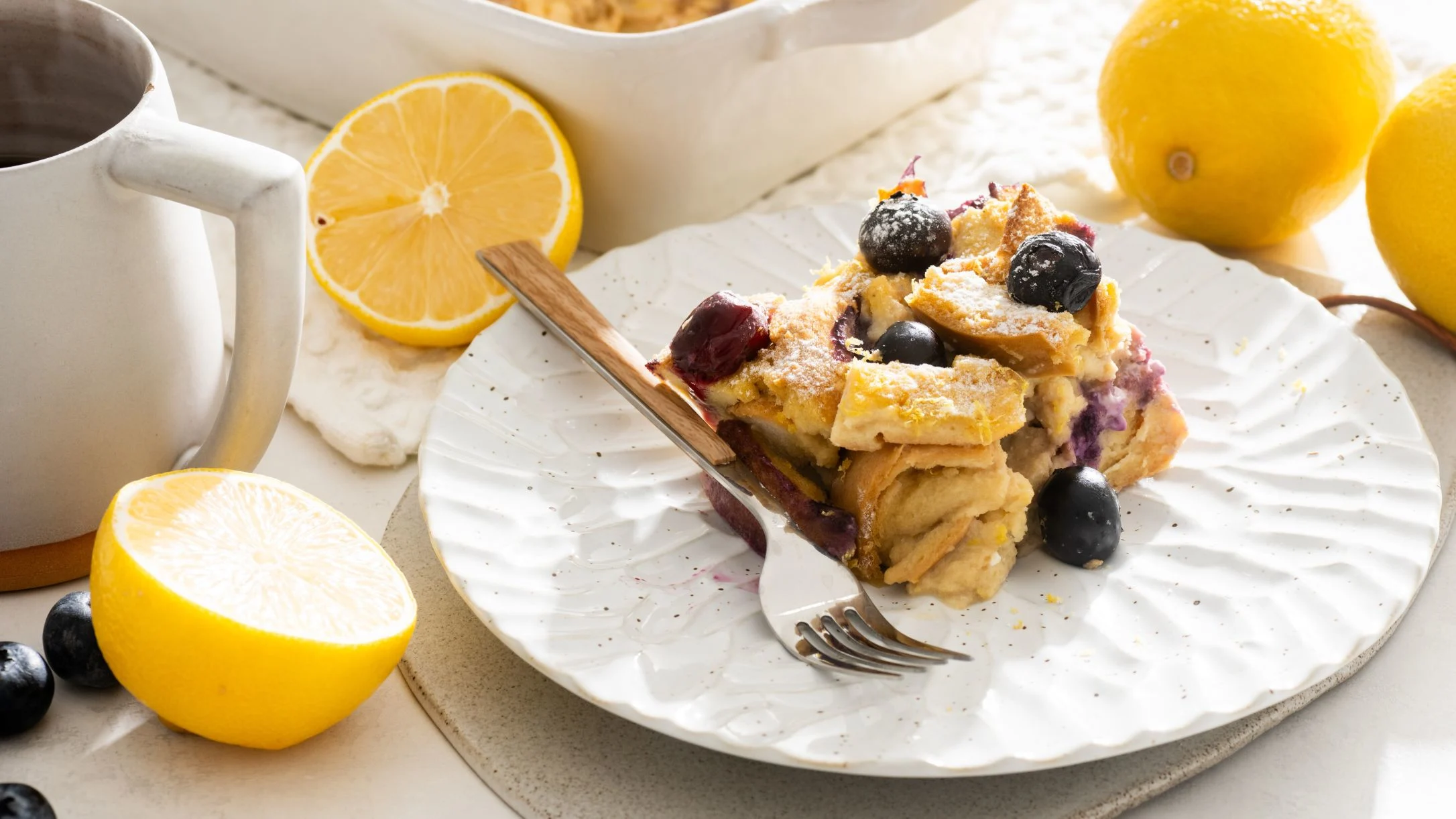 Blueberry bread pudding on white plate with fork, served with coffee and fresh lemons for breakfast.