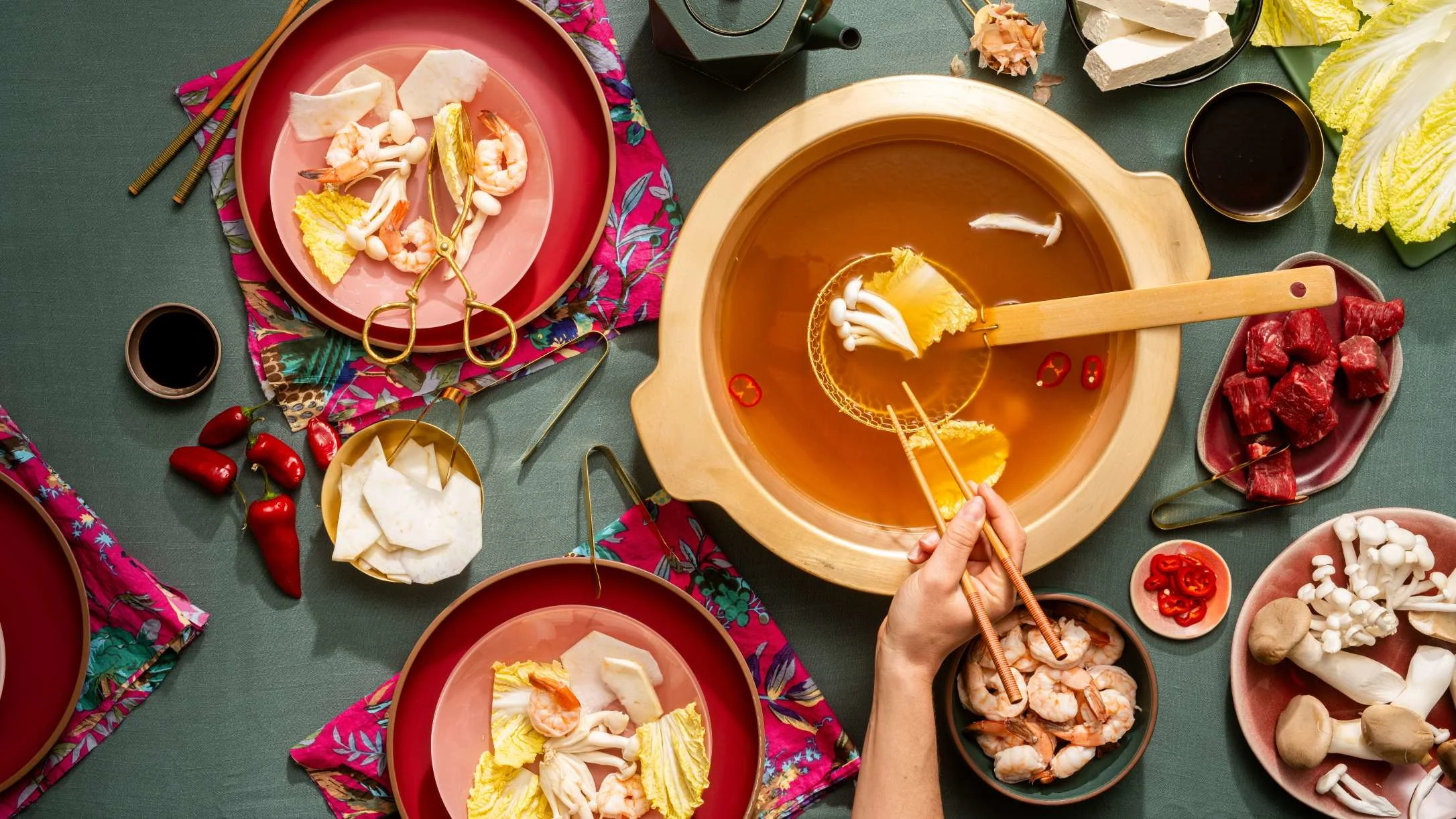 A hand uses chopsticks to cook food in a golden hot pot, surrounded by shrimp, beef, mushrooms, cabbage, and chili peppers.