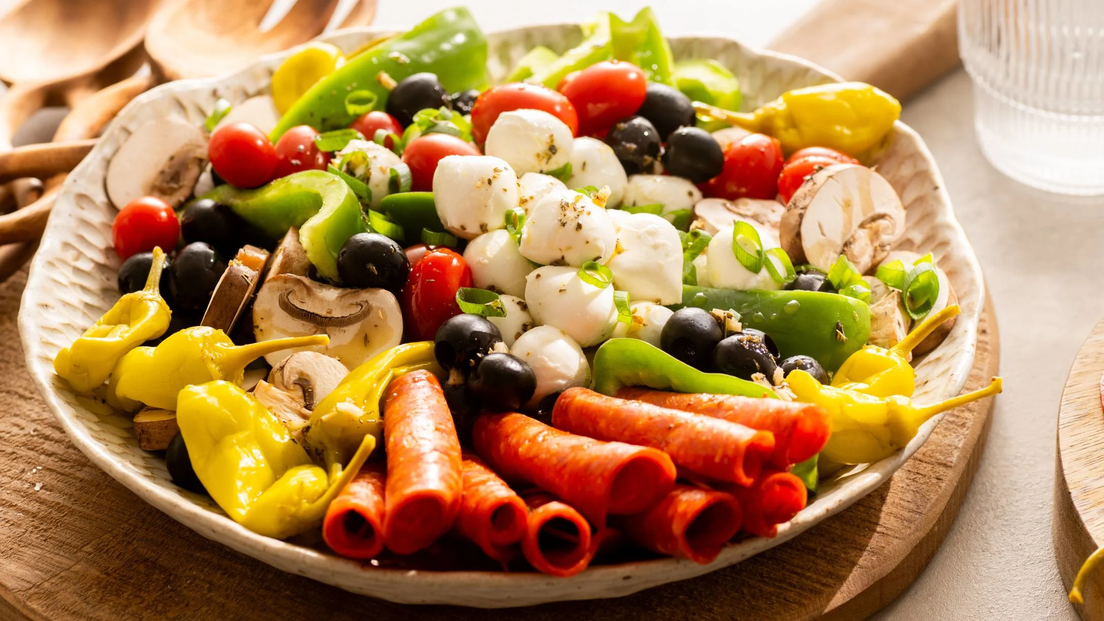 A colorful antipasto platter with rolled pepperoni, mozzarella, olives, cherry tomatoes, peppers, and mushrooms on a ceramic dish.