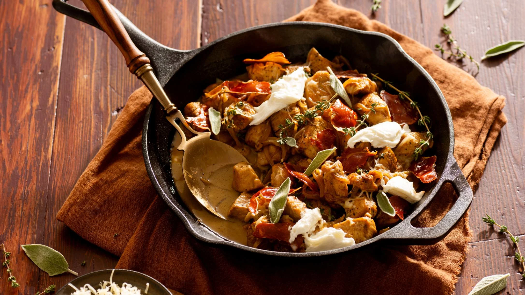 Cast iron skillet with sautéed chicken, prosciutto, fresh herbs, and dollops of burrata cheese on a rustic wood surface.