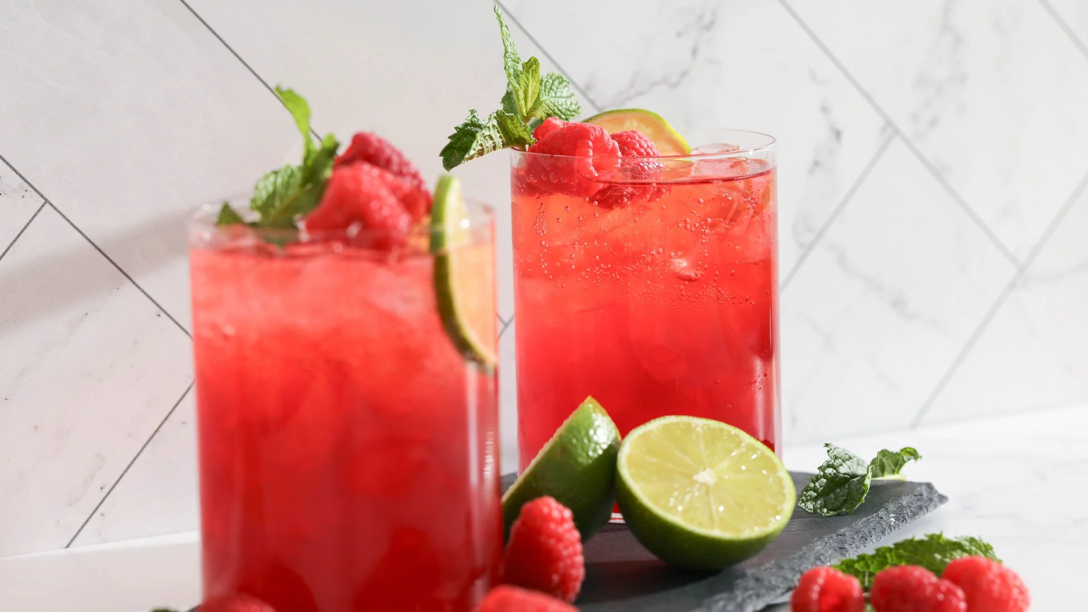 Two tall glasses of red raspberry lime sparkling beverage garnished with mint, lime slices, and fresh raspberries on a slate surface.