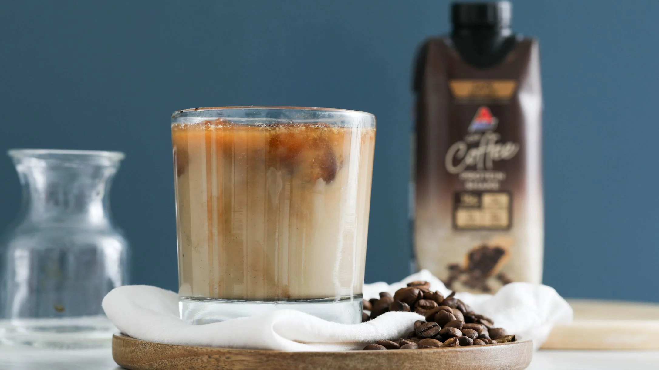Iced coffee in glass with layered effect, coffee beans on wooden plate, bottled coffee drink in background against blue wall.