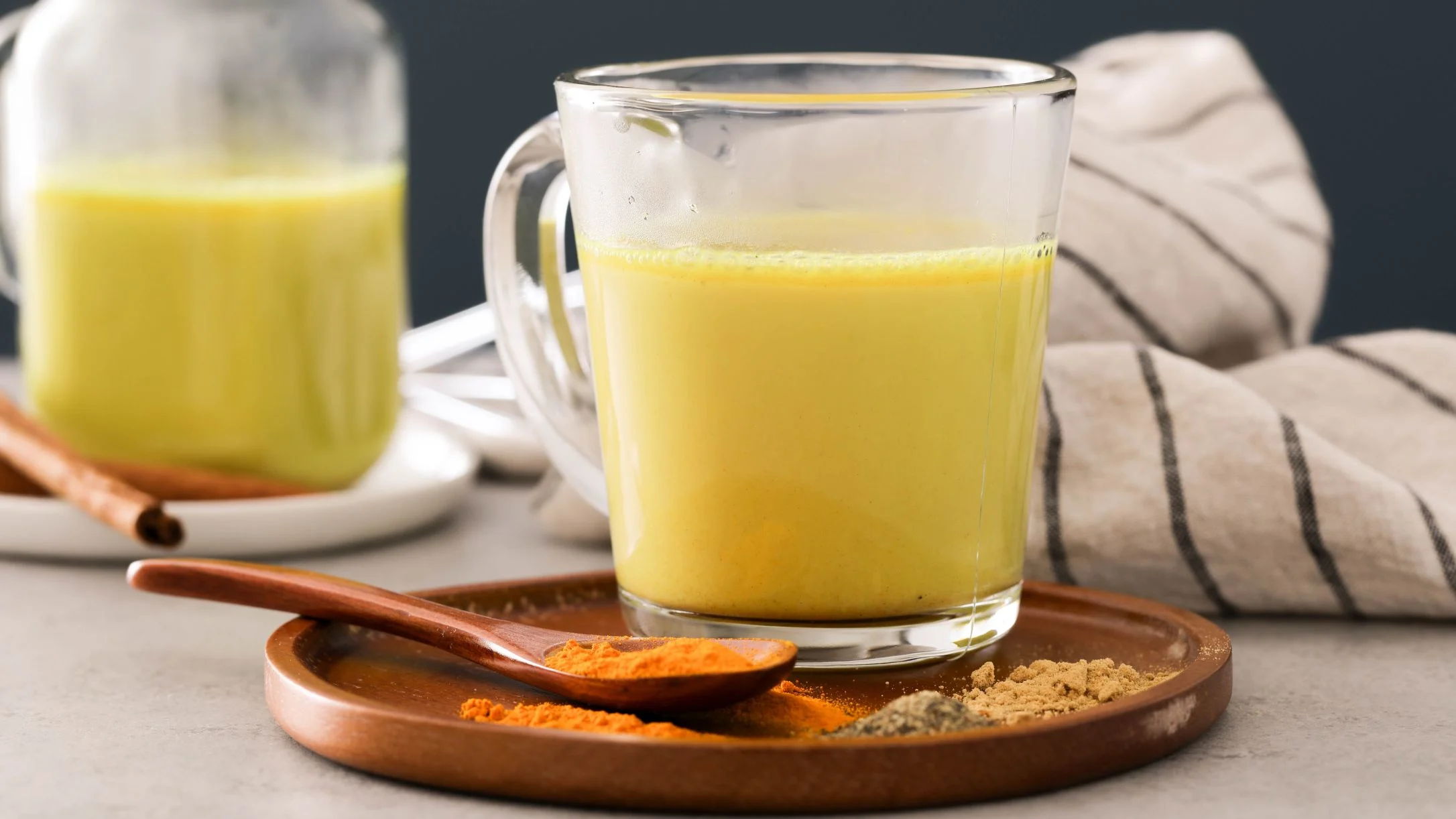 Glass mug of golden turmeric milk with spices on wooden plate, cinnamon stick and striped cloth in background.