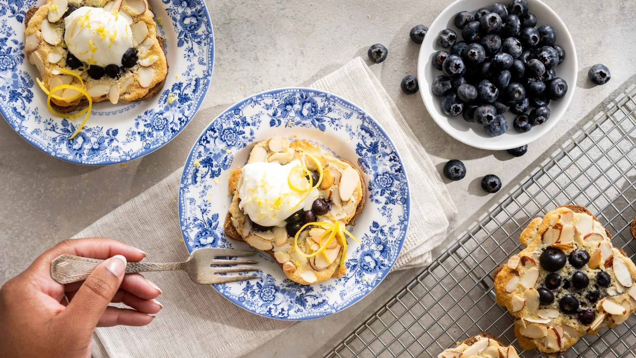 Blueberry almond pastries topped with crème fraîche  and lemon zest on blue floral plates, with a bowl of fresh blueberries nearby.