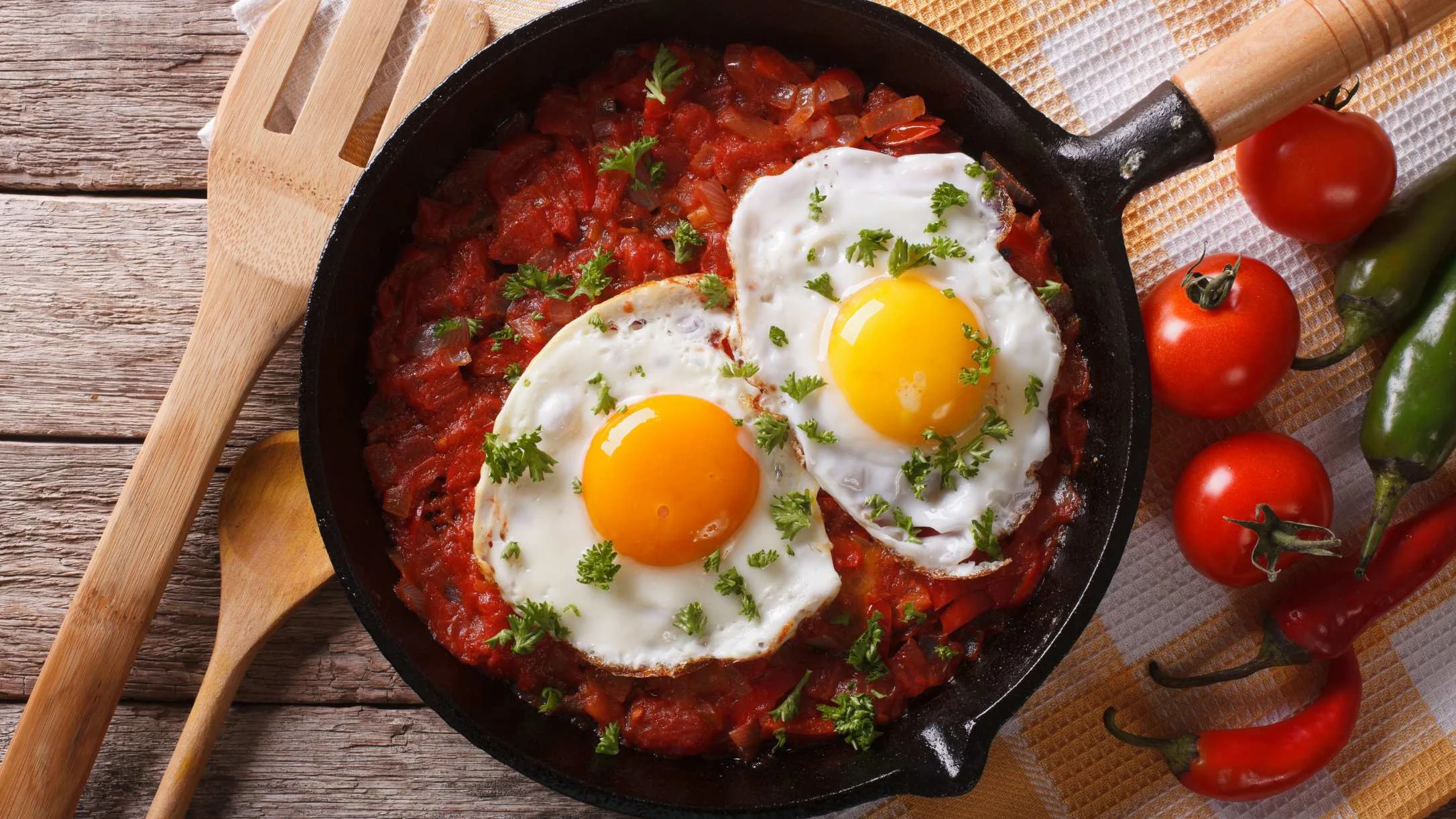 Two sunny-side-up eggs in a cast iron skillet with tomato sauce and parsley, beside fresh tomatoes and green peppers.