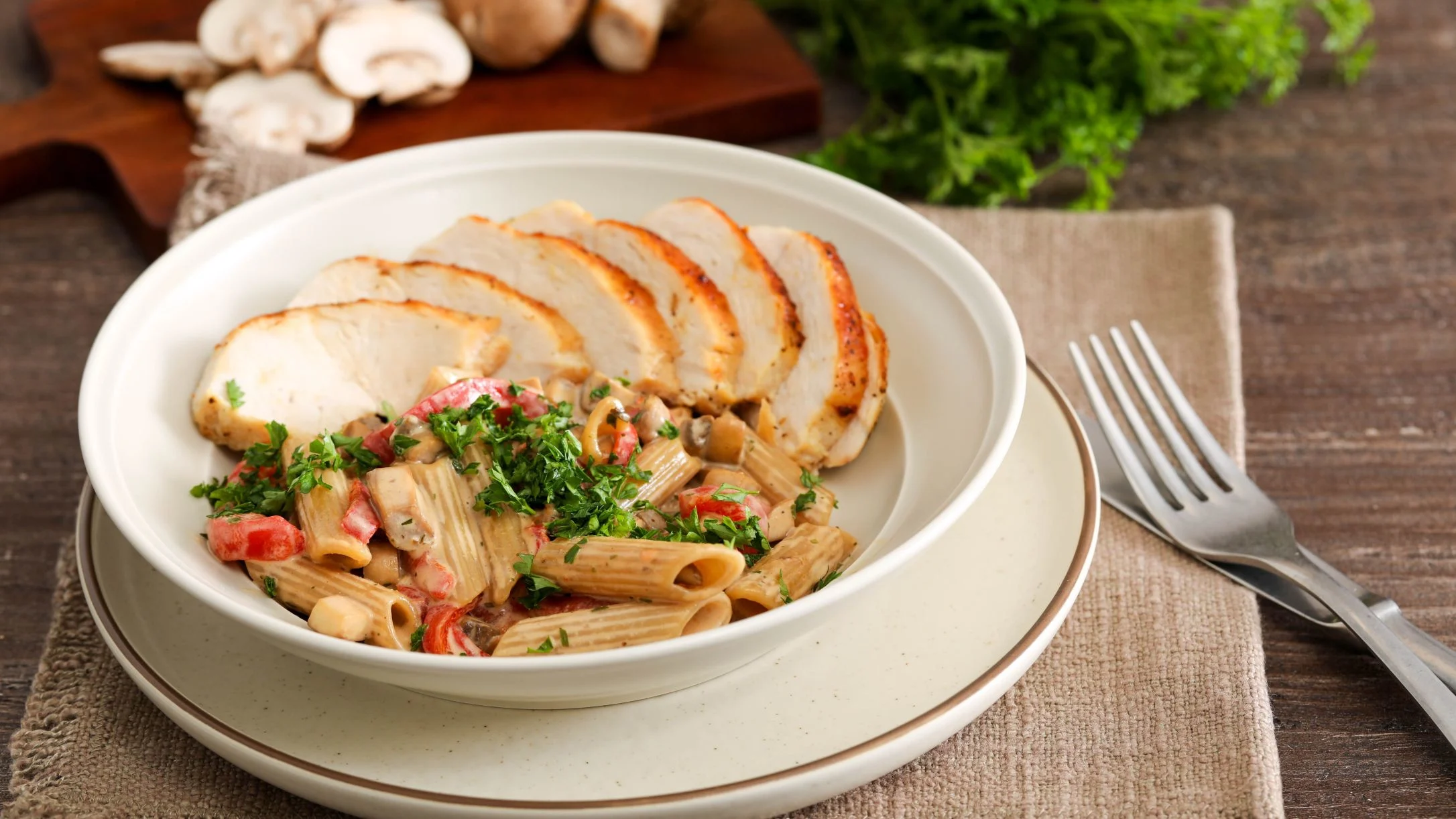 A white bowl with sliced chicken over penne pasta with roasted bell pepper, mushrooms, and fresh parsley, beside a fork.