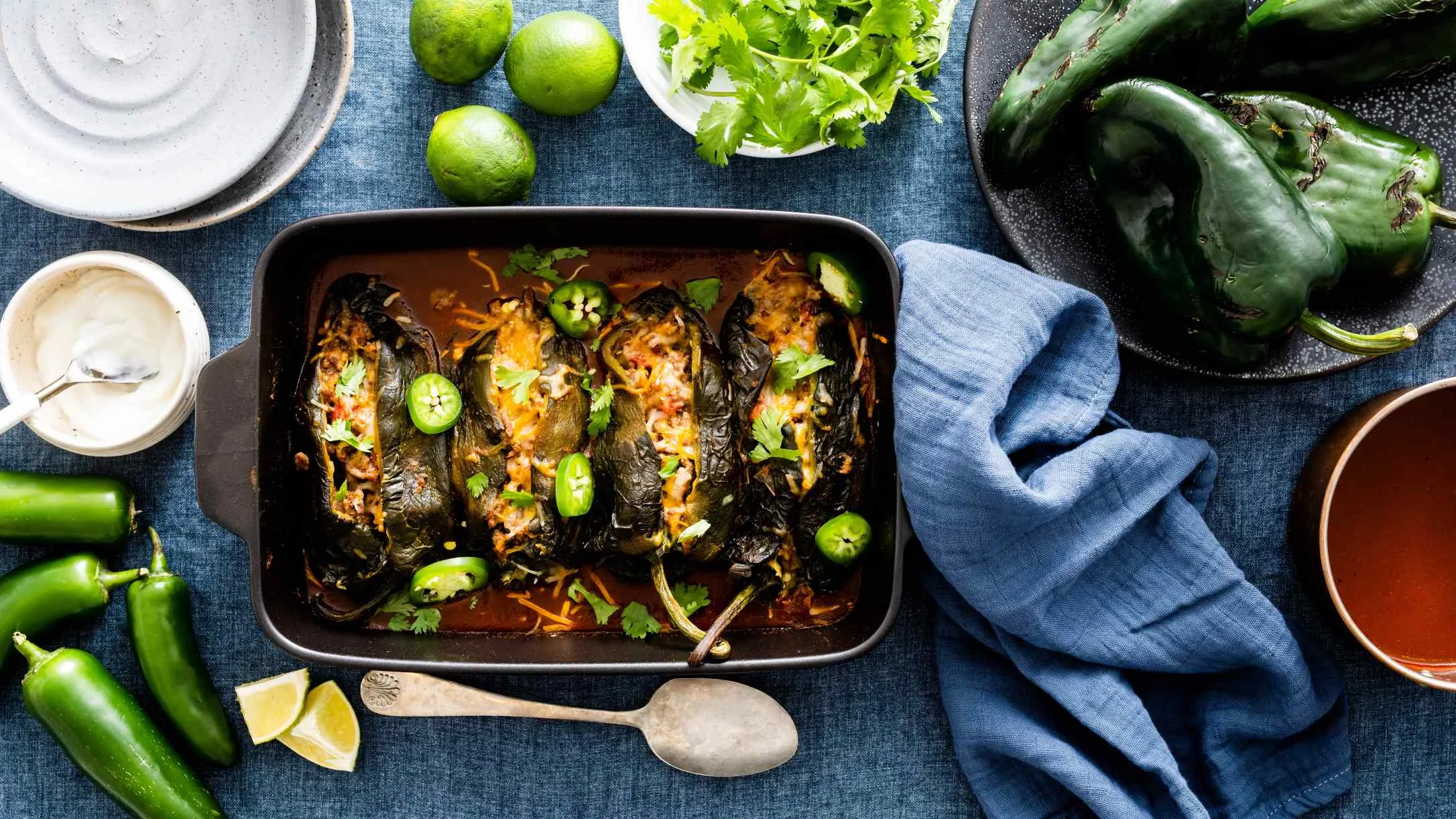 Stuffed poblano peppers in baking dish surrounded by fresh ingredients including limes, cilantro, and jalapeños on blue cloth.