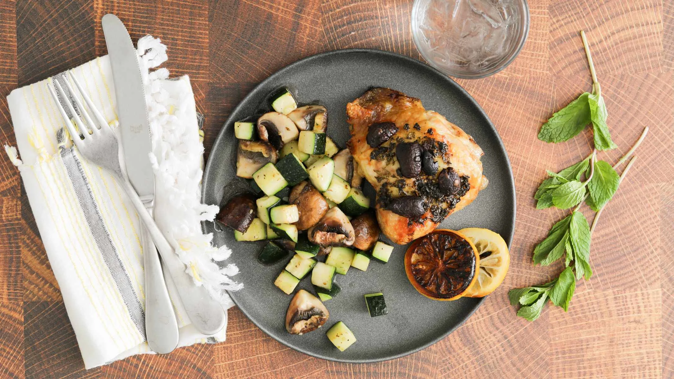 Chicken with mushrooms and zucchini on a dark plate, served with charred lemon and fresh mint on wooden table.