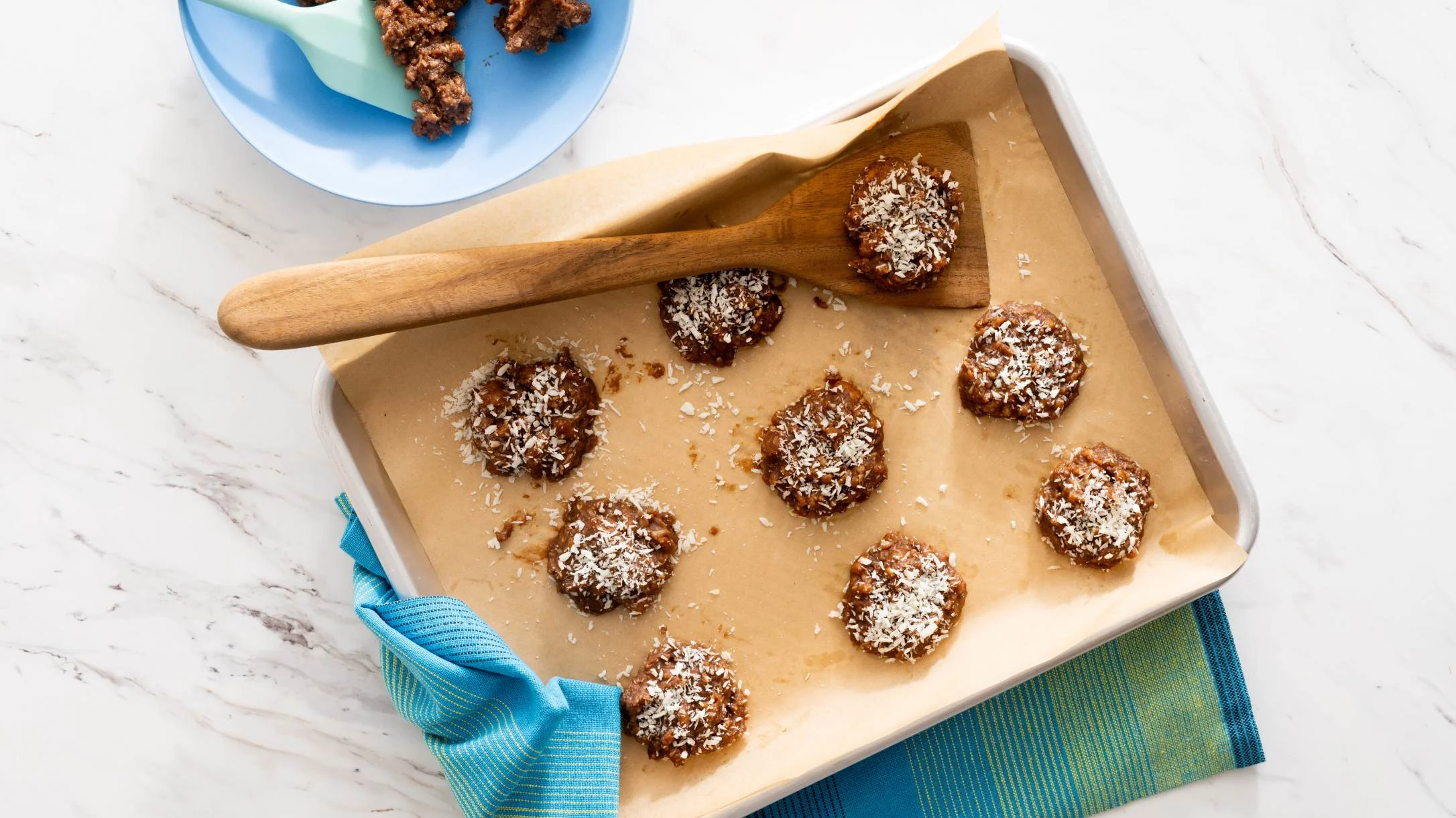 Chocolate coconut cookies on parchment-lined baking sheet with wooden spatula and blue plate of cookie dough nearby.
