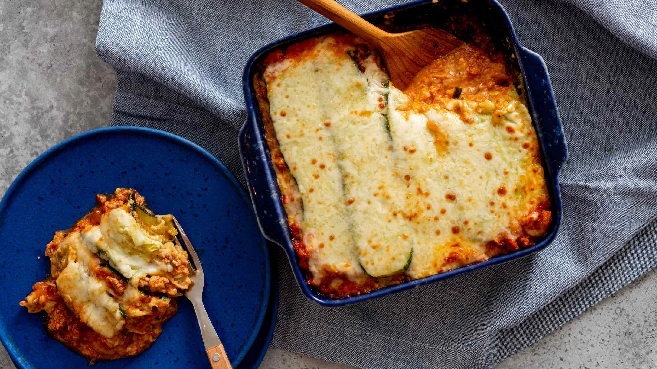 Zucchini lasagna with melted cheese in a blue baking dish, with a serving on a blue plate beside it.