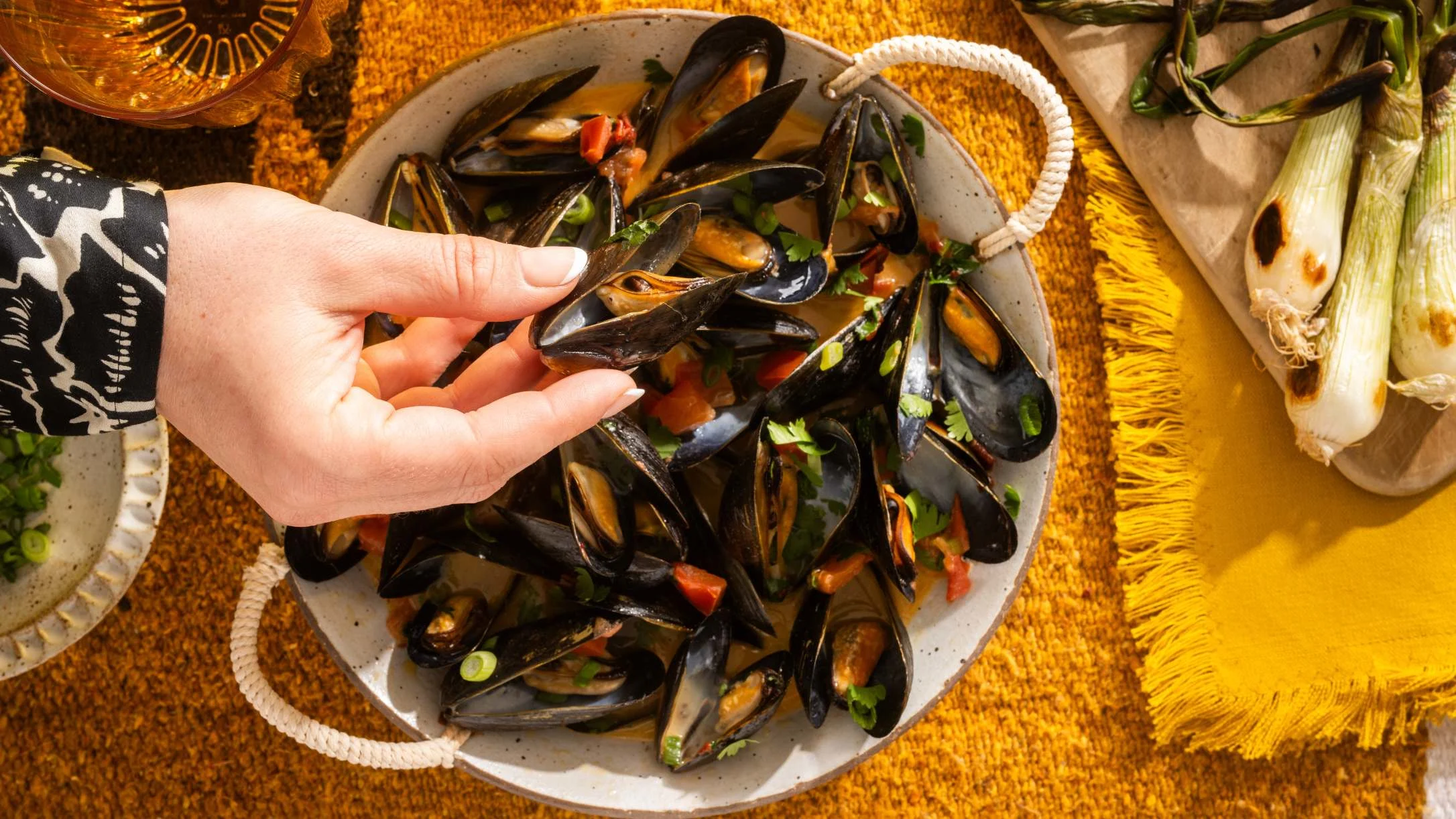 Hand reaching for steamed mussels in a white pot with herbs and tomatoes on a golden yellow textured tablecloth.