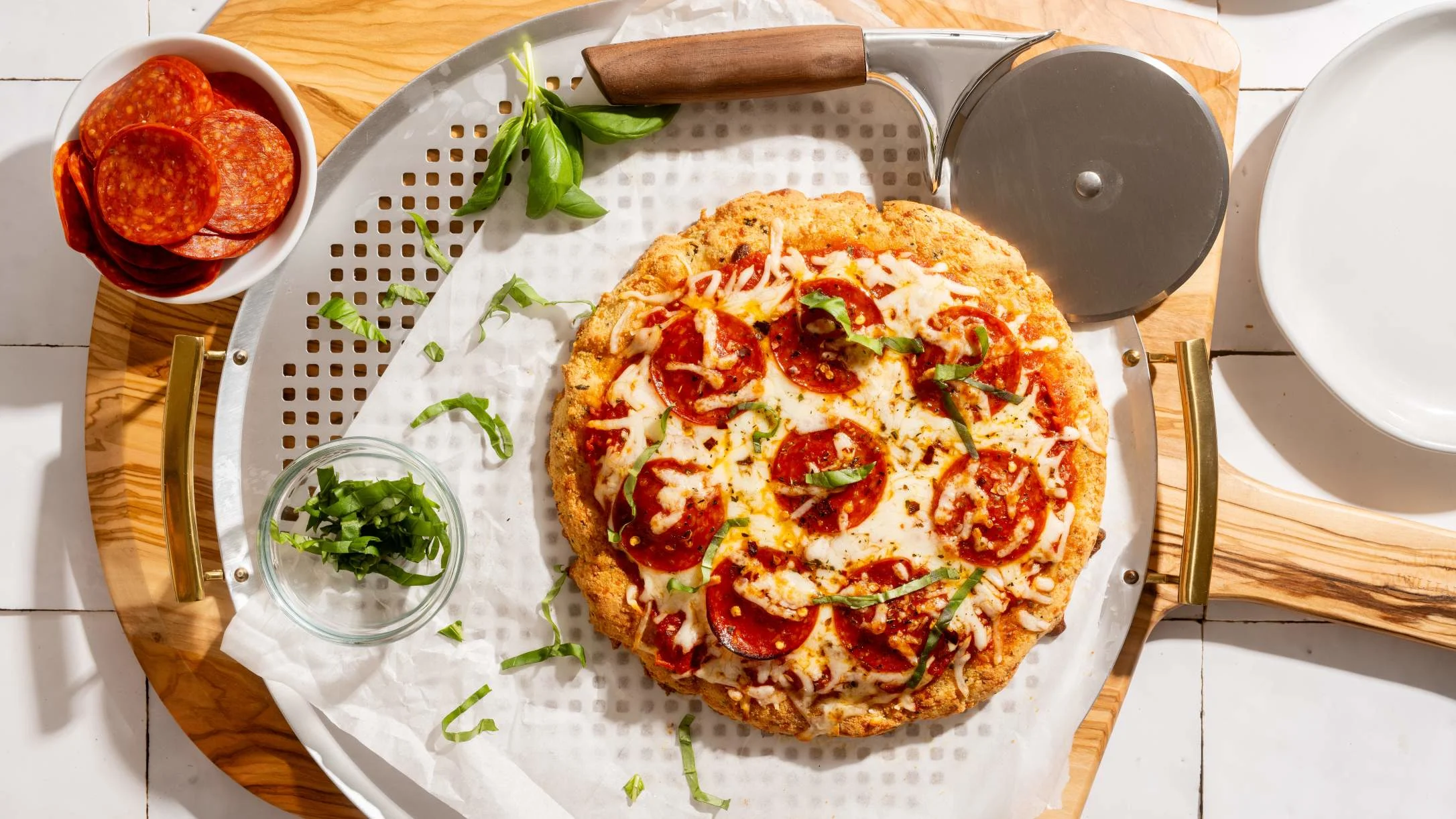 Homemade pepperoni pizza on a round serving tray with fresh basil, chopped herbs, and additional pepperoni slices nearby.