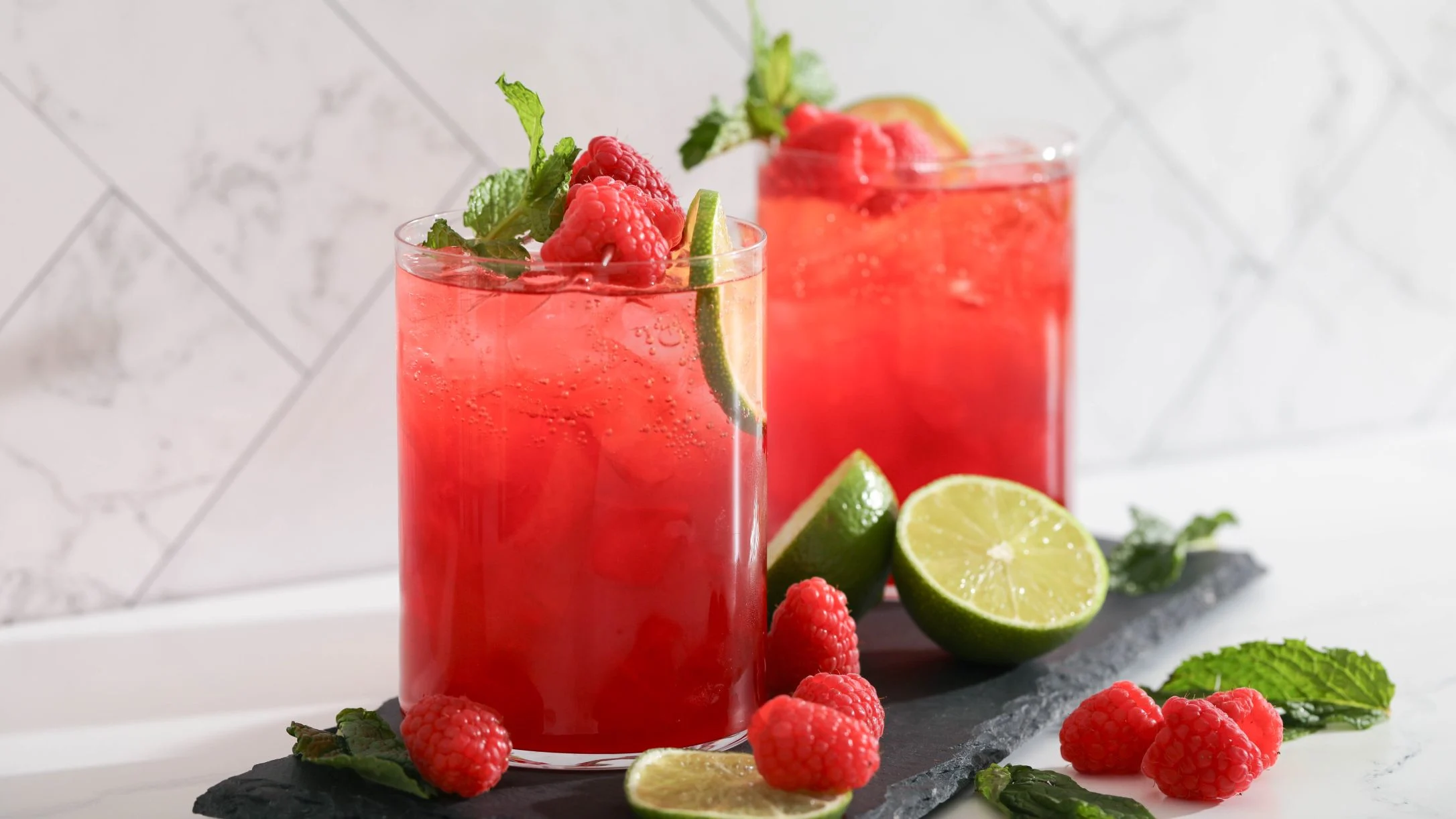 Two red raspberry mocktails garnished with mint and lime, surrounded by fresh raspberries and lime slices on a slate board.
