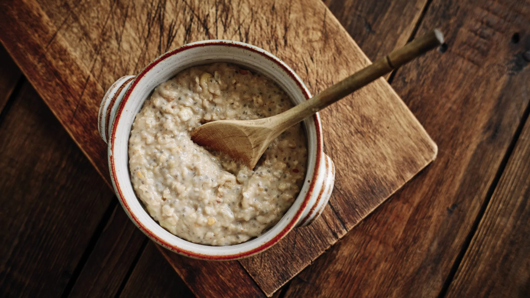 A white ceramic bowl with red trim filled with creamy porridge, topped with a wooden spoon, resting on a rustic wooden board.