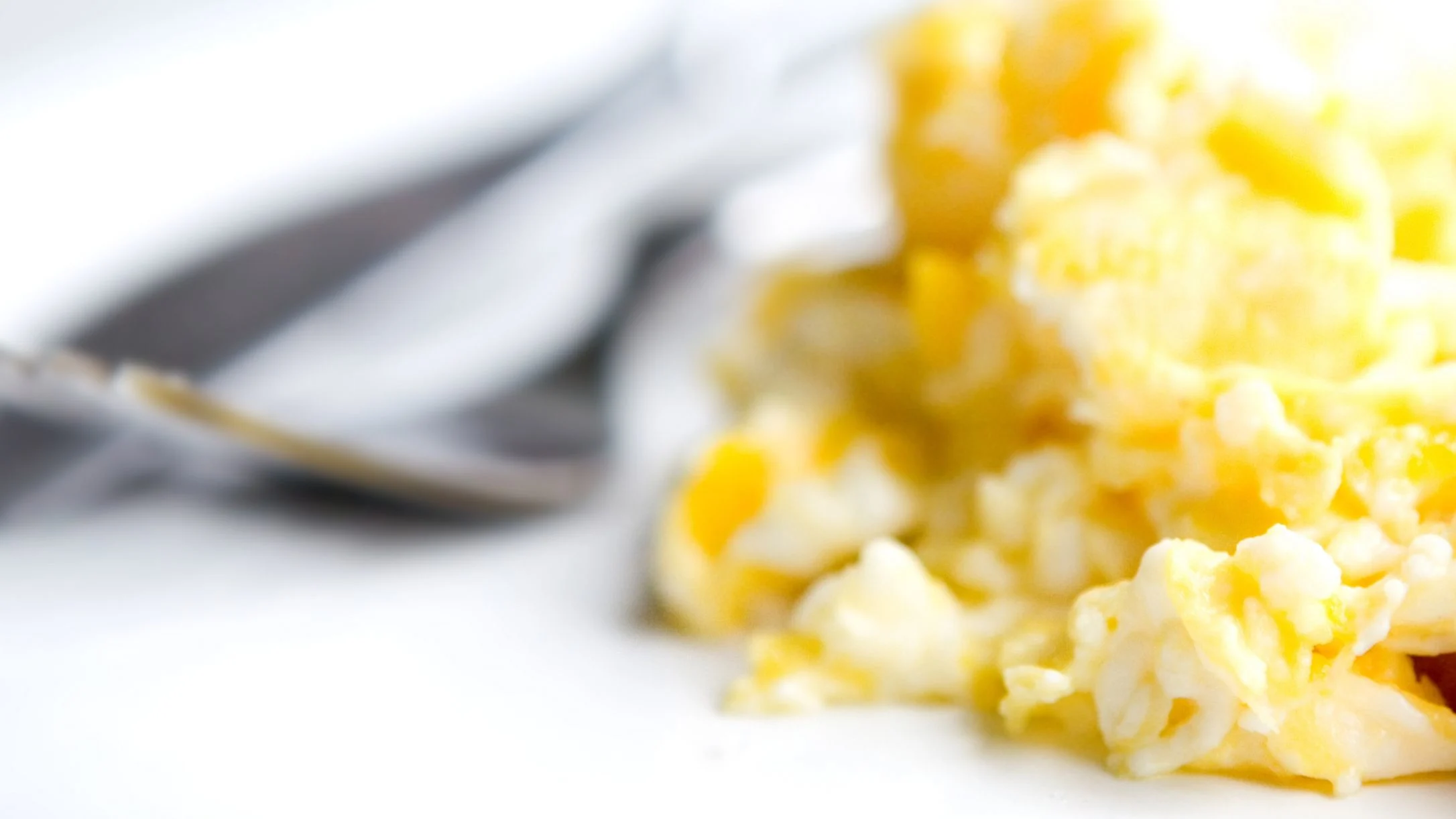 Close-up of fluffy yellow scrambled eggs on a white plate with blurred silverware and gray napkin in the background.