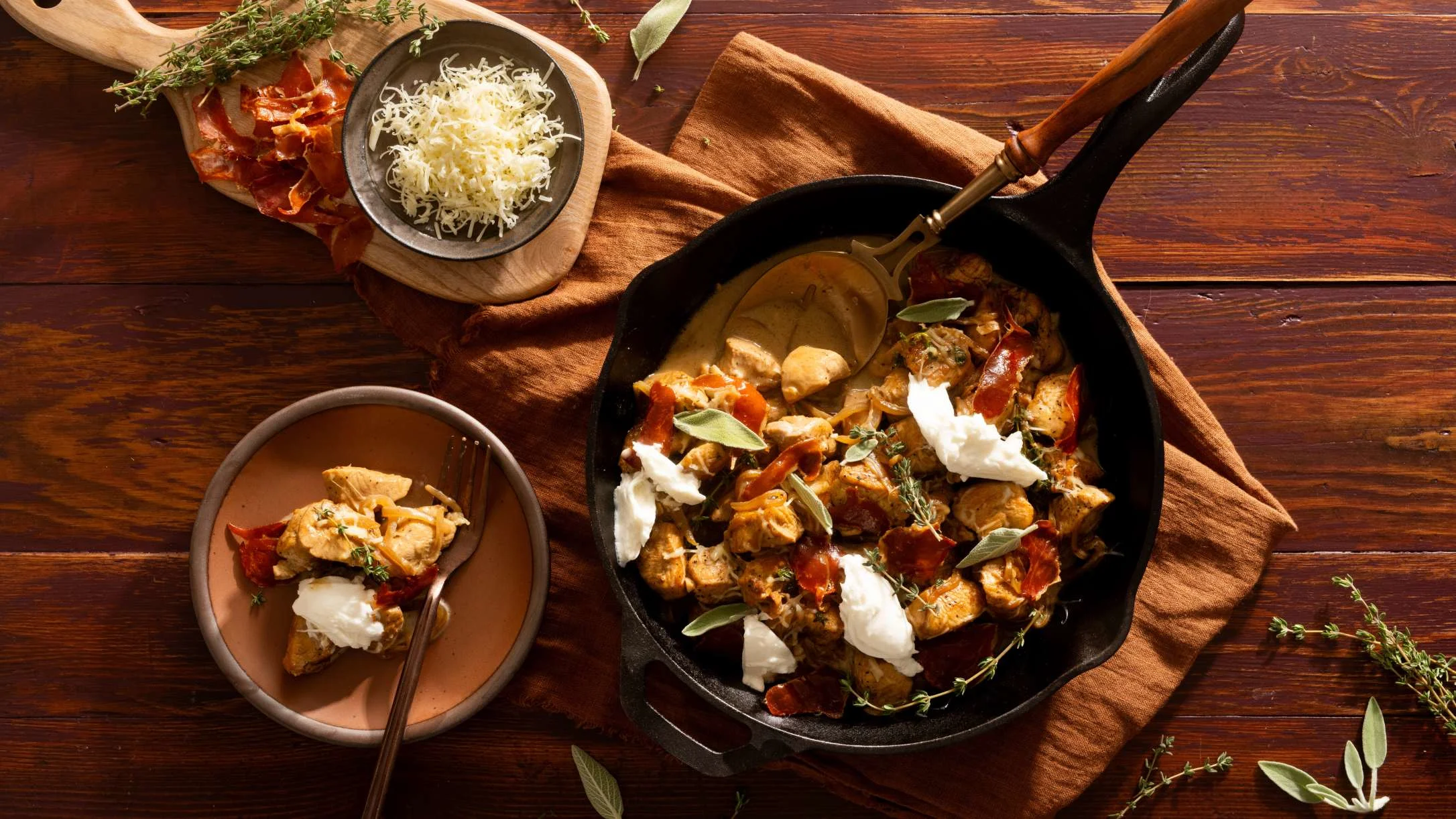 Cast iron skillet with chicken, prosciutto, and burrata cheese, served in a bowl, with shredded cheese and herbs on a wooden table.