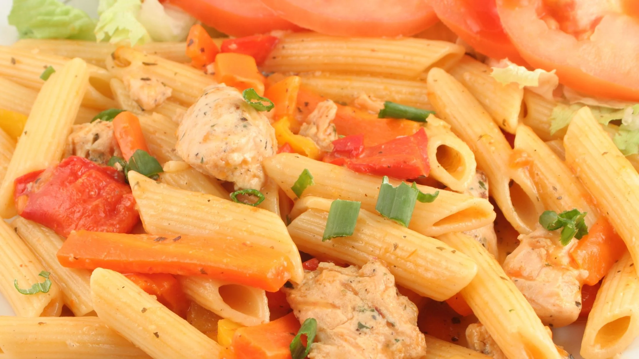 Penne pasta dish with chicken, red and orange bell peppers, carrots, and green onions, garnished with fresh tomato slices.