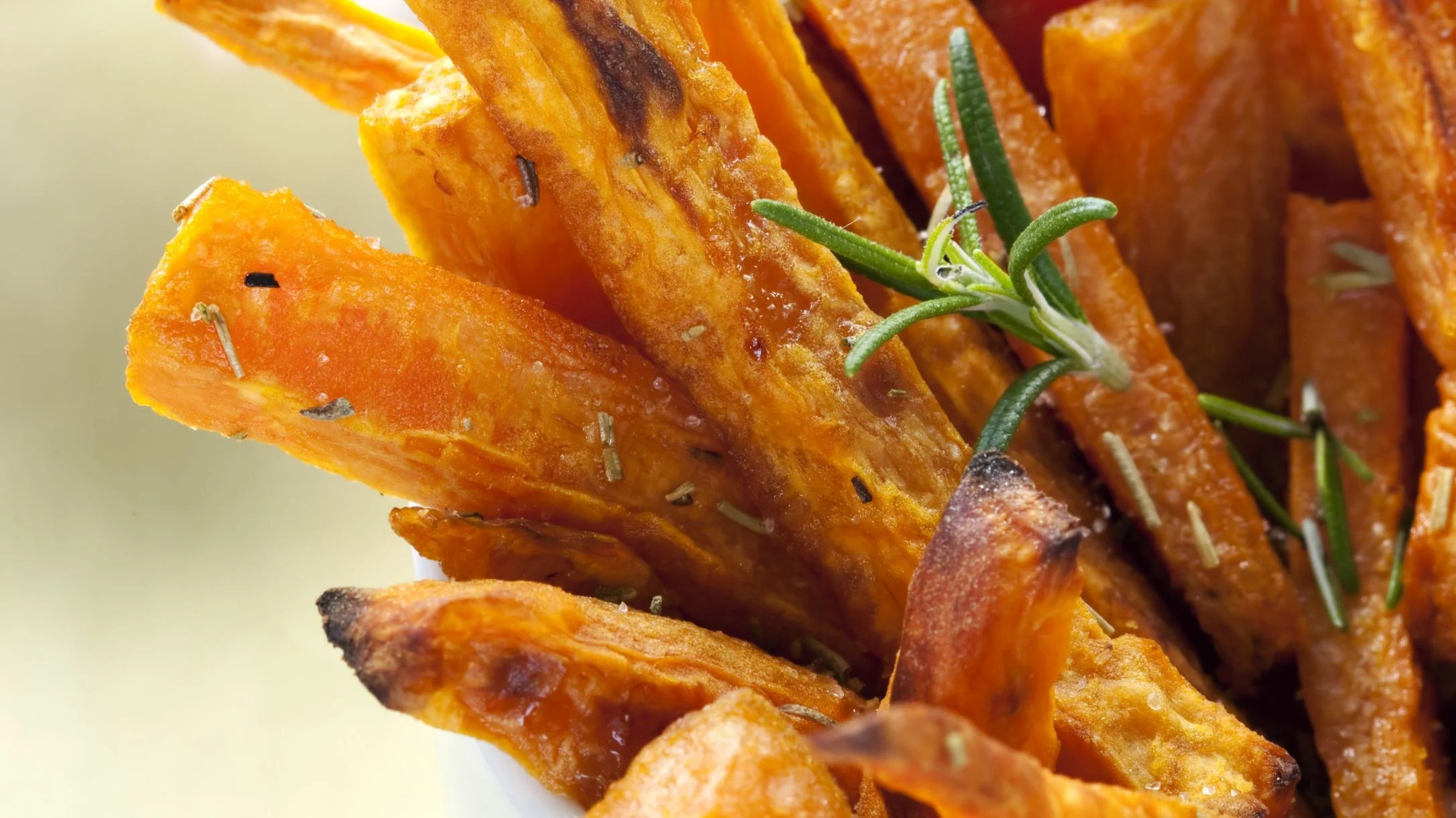 Close-up of golden-orange roasted sweet potato fries seasoned with rosemary sprigs and salt on a white plate.