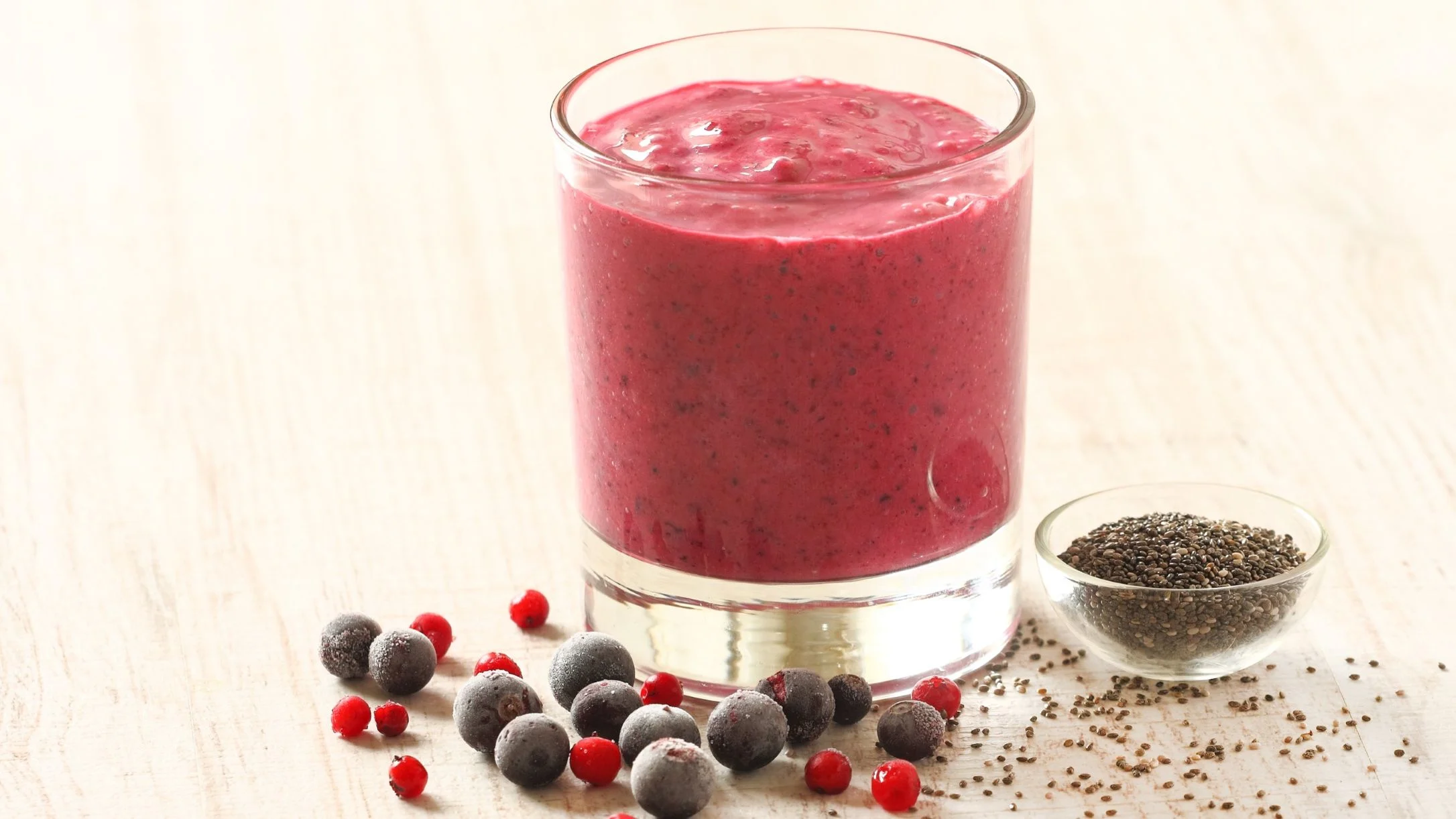 A vibrant red berry smoothie in a glass, surrounded by frozen berries and a small bowl of chia seeds on a light wooden surface.