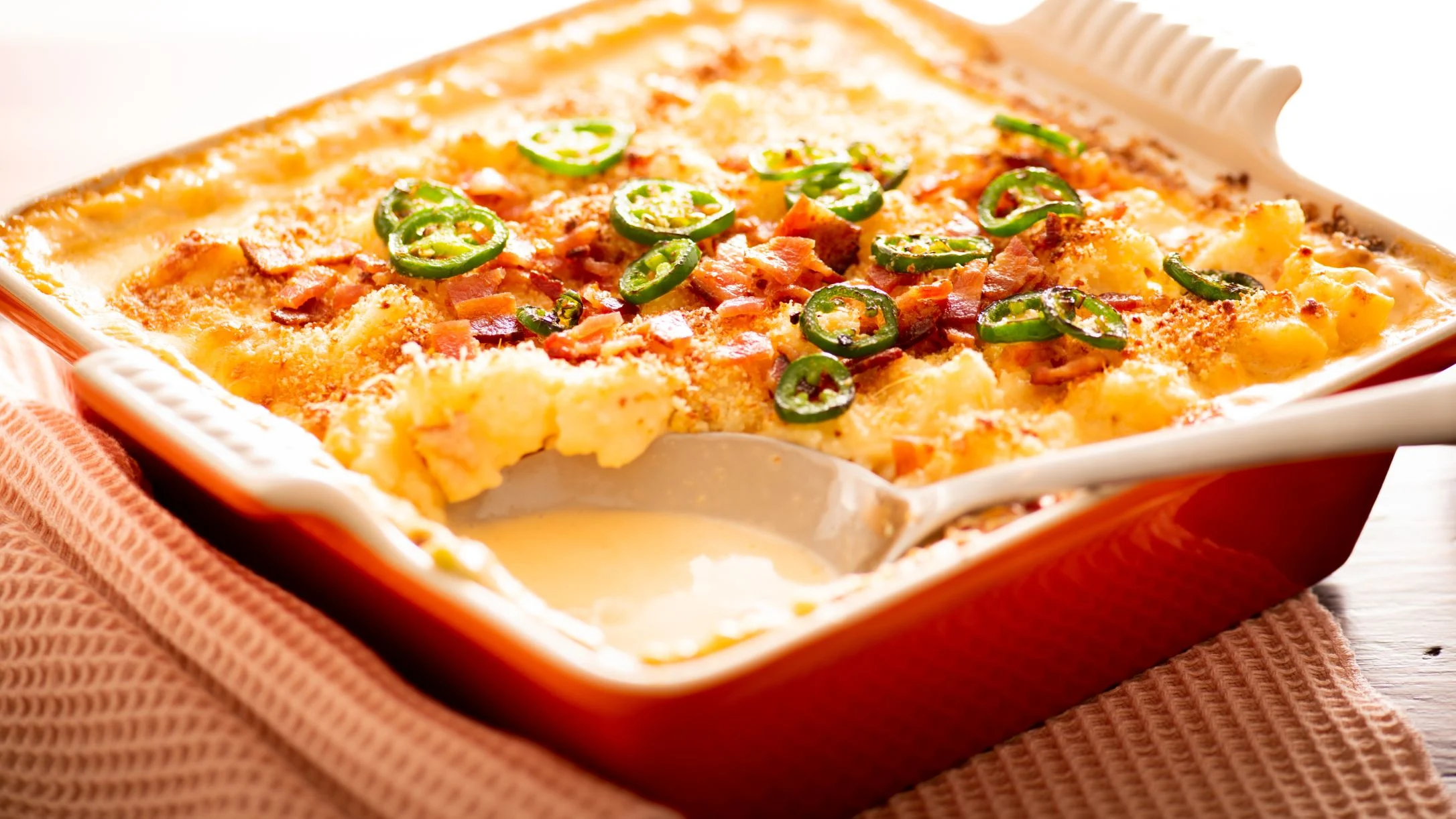 Baked mac and cheese casserole in orange dish topped with bacon bits and sliced jalapeños, with one serving removed.