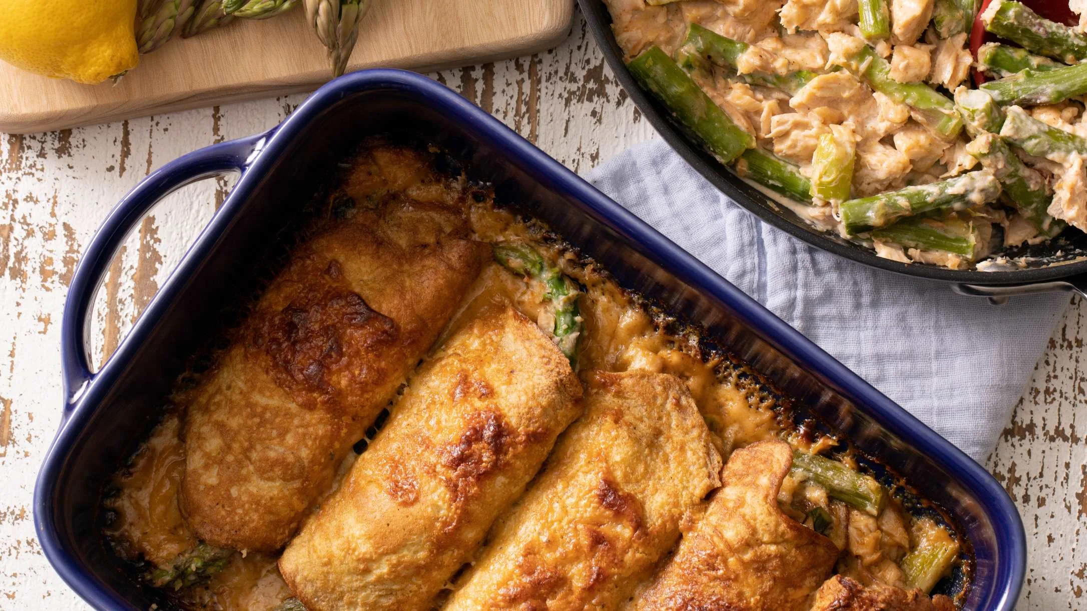 Golden-baked filled crepes in a blue baking dish, beside a skillet with salmon and asparagus filling, and a lemon on a cutting board.