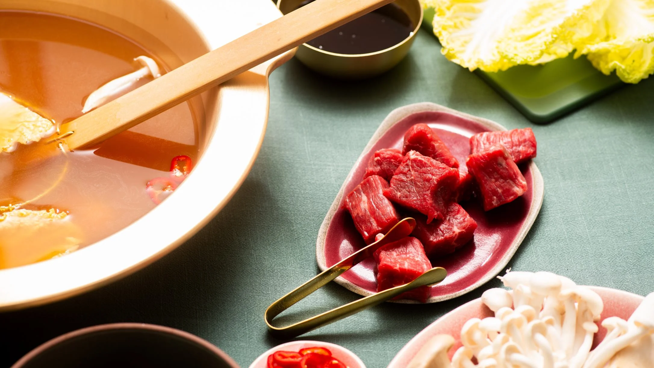 Hot pot setup with raw beef chunks on a pink plate, mushrooms, cabbage, dipping sauce, and a broth-filled pot with tongs.