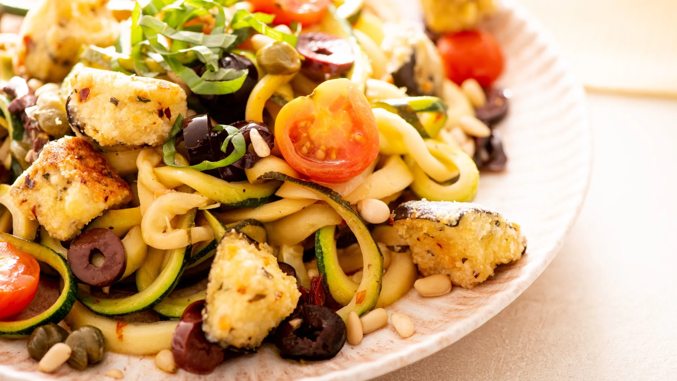 Pasta dish with zucchini noodles, cherry tomatoes, olives, eggplant cubes, and herbs served on a white plate.