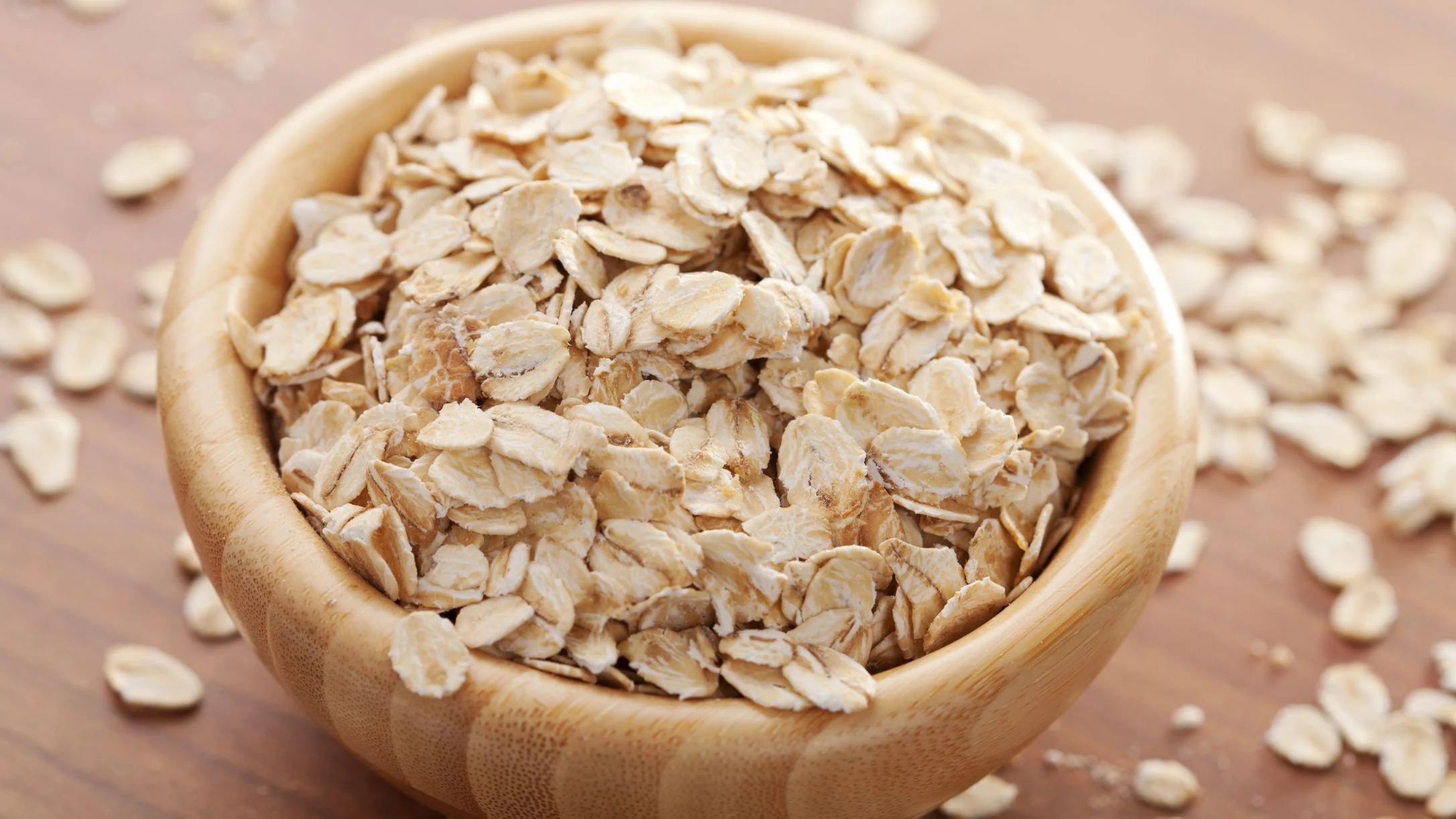 A wooden bowl filled with dry rolled oats, with scattered oat flakes on a brown wooden surface.