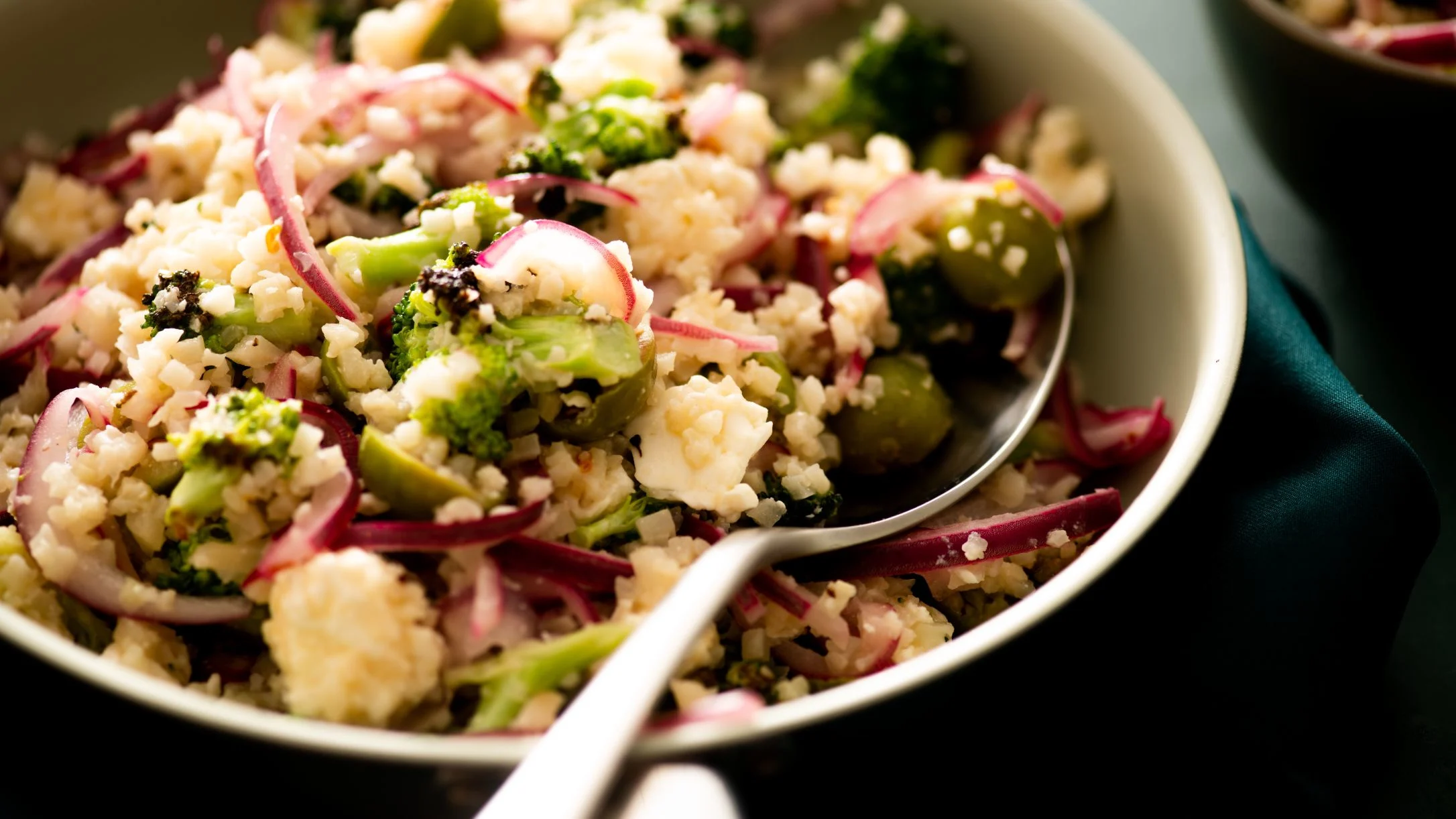 Skillet with cauliflower rice, broccoli, red onion slices, and cheese with serving spoon.