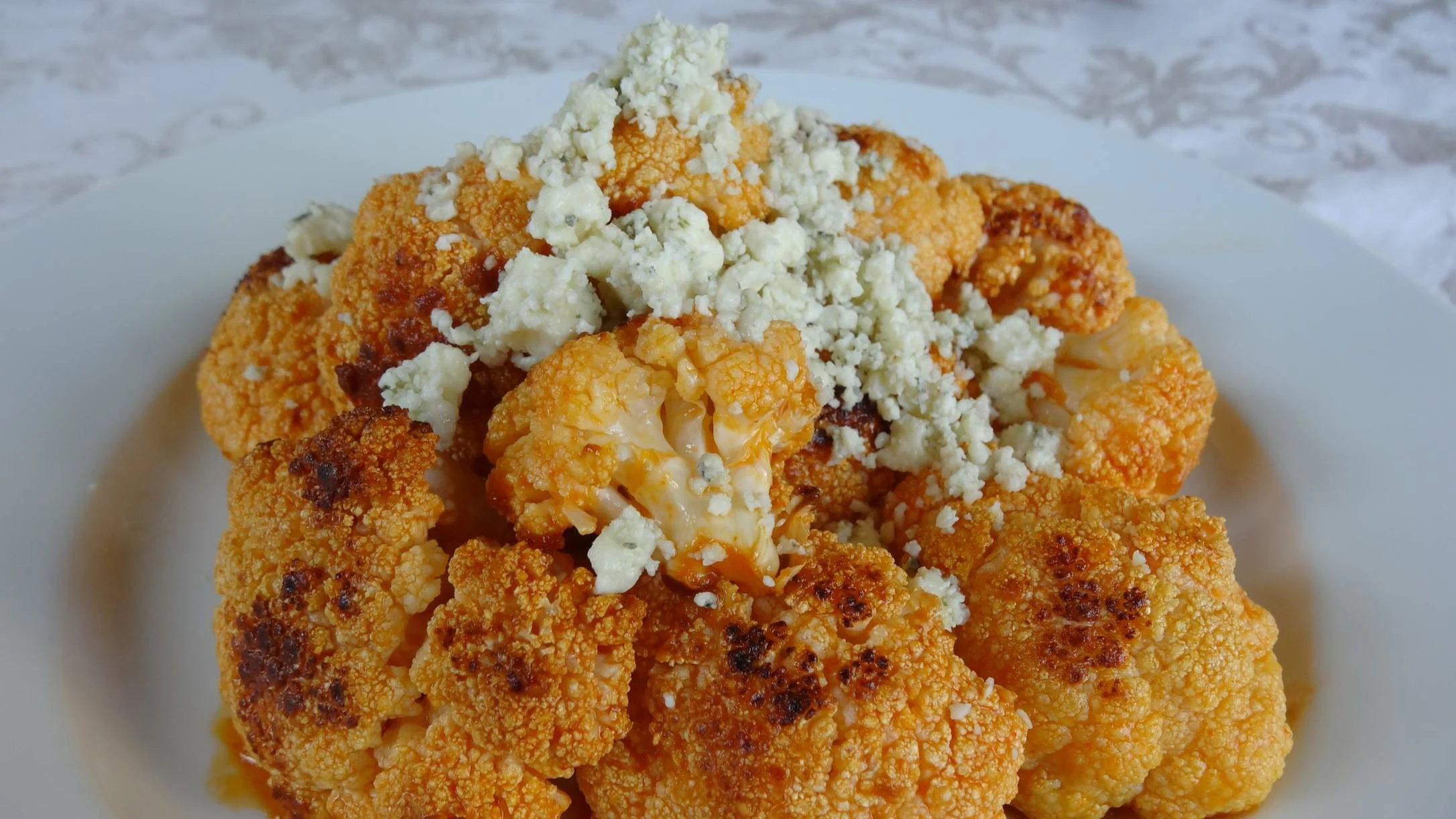 Roasted buffalo cauliflower coated in orange sauce, topped with crumbled blue cheese, served on a white plate.