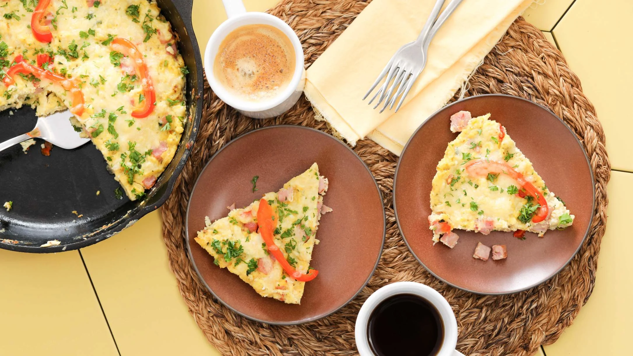 Two plates with frittata slices topped with red peppers and herbs, beside a cast iron skillet and two cups of coffee.