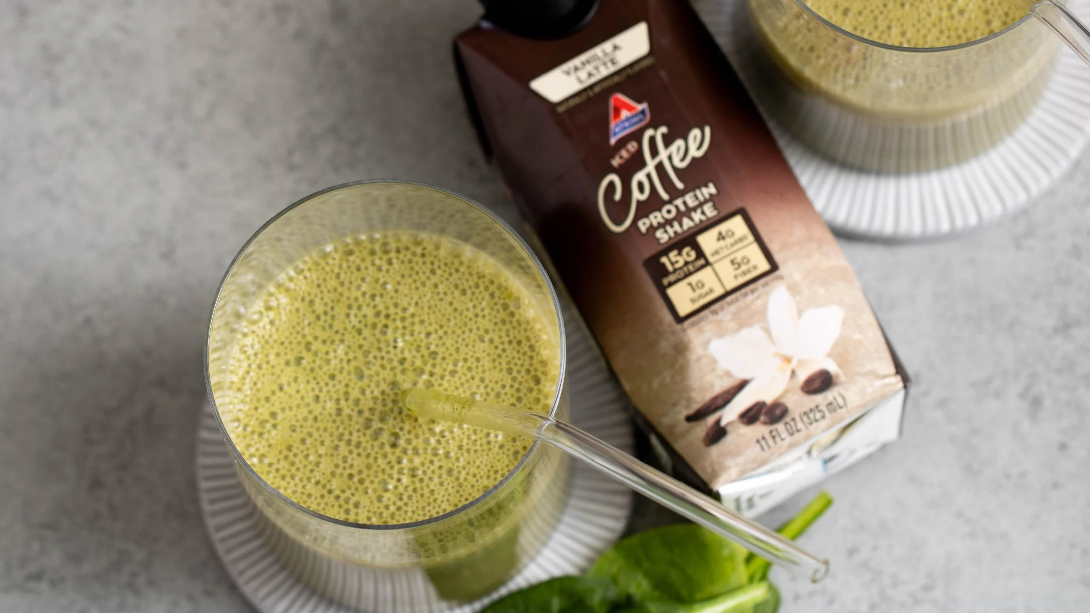 A green smoothie with a glass straw next to an Atkins Vanilla Latte Iced Coffee Protein Shake carton and fresh spinach leaves.
