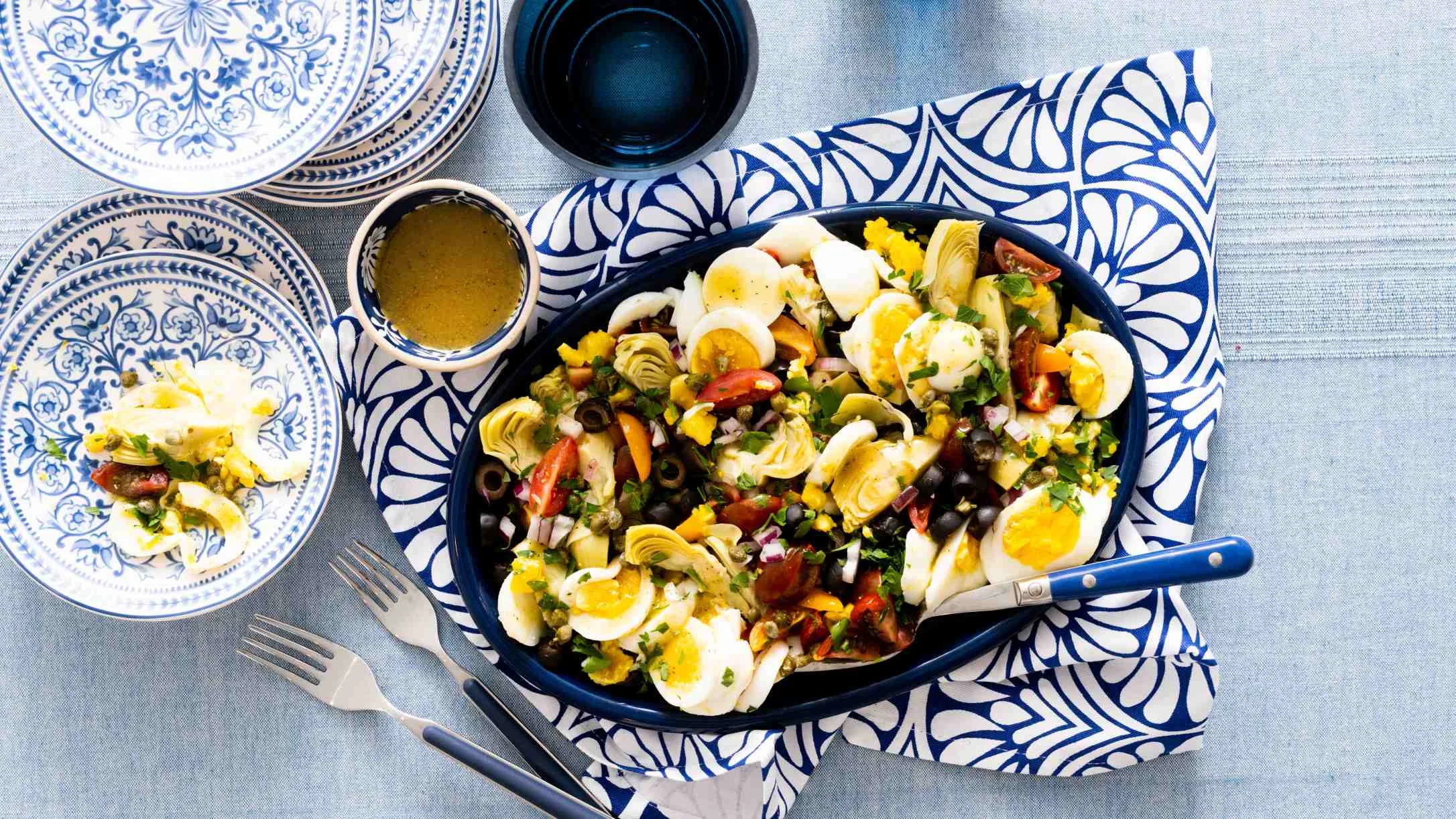 Colorful Mediterranean salad with eggs, artichokes, and olives in a blue bowl, served with blue and white patterned dishes.