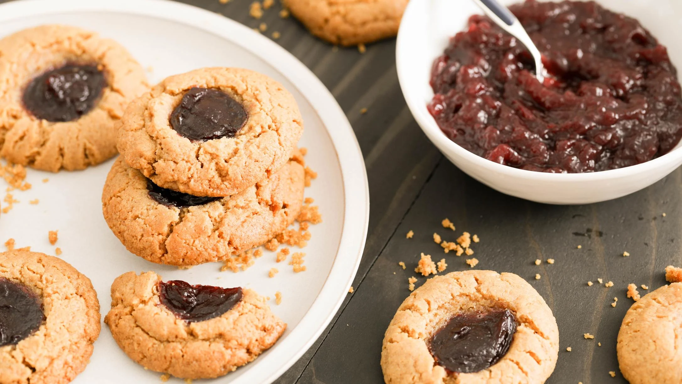 Thumbprint cookies with berry jam centers on a white plate beside a bowl of dark berry preserves on a black surface.